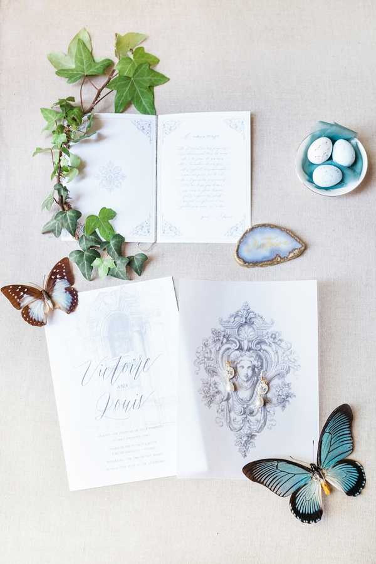 A flat lay detail shot of a French wedding stationery suite for 'Victoire and Louis,' displayed on a linen-toned surface. The invitation suite features white and pale grey card stock with ornate baroque-style illustrated engravings, including a classical female figure motif, and calligraphic script lettering. Styling props arranged around the stationery include a trailing ivy vine, two taxidermy or decorative butterflies — one brown and white, one teal and black — a small agate slice in blue-grey tones, and a pale blue dish holding three small speckled white eggs. The overall palette is soft white, pale grey, dusty teal, and natural green, suggesting a refined classical French aesthetic.