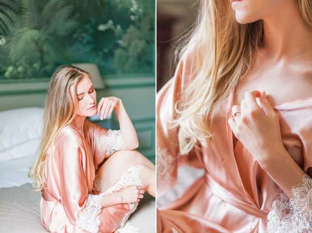 A bridal getting-ready scene presented as a two-image diptych. On the left, the bride sits on a white bed wearing a blush/rose gold satin robe over white lace lingerie, with long blonde hair loose, eyes closed, in a relaxed posed portrait with lush green foliage visible through a window in the background. On the right, a close-up detail shot focuses on the bride's neckline and hand, showcasing the blush satin robe fabric, white lace trim underneath, and what appears to be an engagement ring on her finger. Both shots are softly lit with warm natural light and have a light, airy film-style aesthetic. The composition combines a three-quarter portrait with an intimate detail close-up.