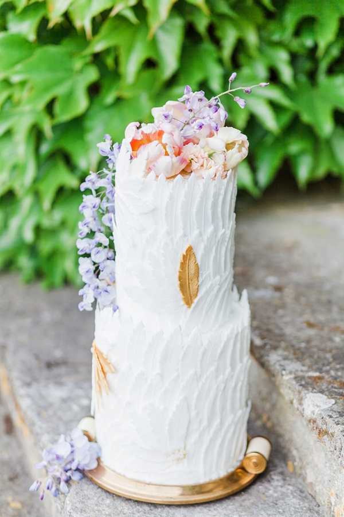 Close-up detail shot of a two-tier wedding cake placed on a stone surface outdoors. The cake features textured white buttercream with a raked or feathered finish, accented with gold leaf decorations on both tiers, and sits on a gold cake board. The top is adorned with fresh flowers including blush and coral peonies, lavender wisteria sprigs, and white blooms, with additional wisteria cascading down the left side of the cake. The overall palette combines white, gold, blush, coral, and soft lavender, giving the design a romantic garden-inspired aesthetic.