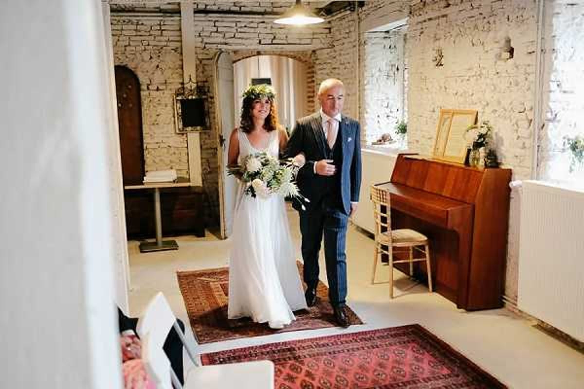 The bride is walking down the aisle during an indoor ceremony, escorted by a male companion — likely her father — who is dressed in a navy blue suit with a pink tie. The bride wears a flowing white sleeveless dress with a relaxed, bohemian silhouette and carries a large bouquet of white blooms and greenery; she also wears a green floral crown on her curly hair. The venue has a rustic, converted-barn aesthetic with whitewashed exposed brick walls, a patterned red and navy runner rug on a pale floor, a wooden upright piano to the right, and white chiavari chairs visible at the edges of the frame. The shot is taken from a medium-wide angle from behind the seated guests, capturing the couple mid-procession in natural window light.