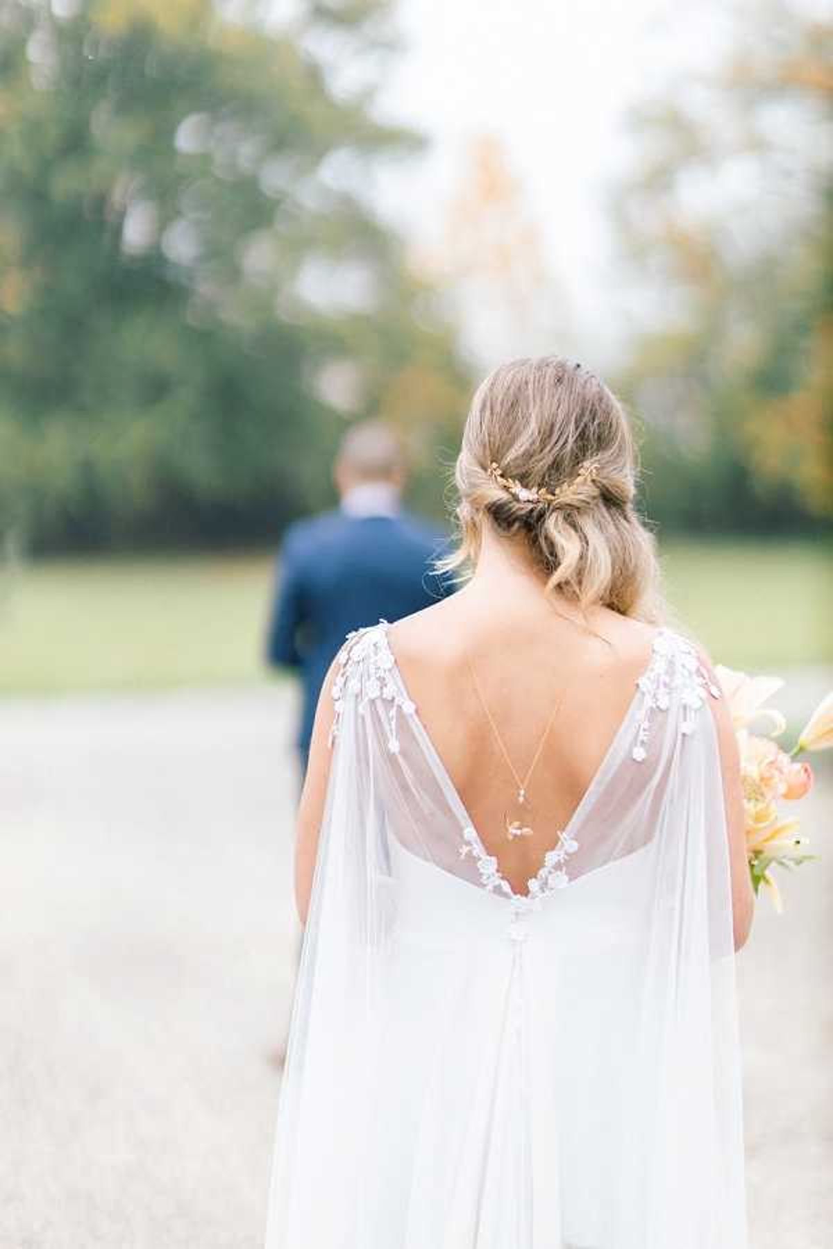 A close-up portrait taken from behind the bride, focusing on the back detail of her white wedding dress, which features a deep V-back, floral appliqué along the shoulder straps, and a flowing chiffon cape overlay. She wears a delicate back necklace with a small pendant and a gold floral hair accessory in her loosely pinned updo. She holds a bouquet with peach and coral blooms visible at the edge of the frame. The groom, dressed in a navy suit, is visible out of focus in the background walking along a gravel path, set against a soft outdoor garden or parkland setting. The overall styling is romantic and boho-influenced, with soft natural light and a muted, airy palette.