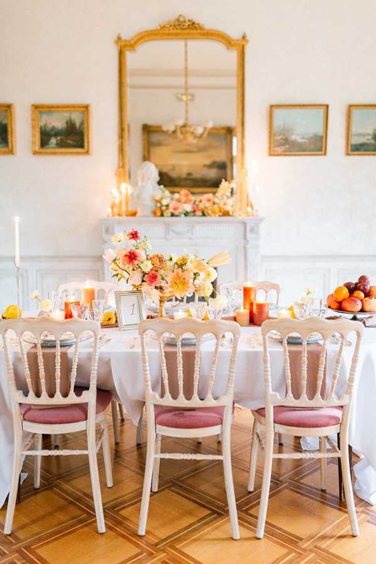 A styled reception tablescape shot indoors in what appears to be a chateau salon, viewed from a slightly elevated angle. The rectangular dining table is dressed in a white linen tablecloth and set with crystal glassware, gray charger plates, and a small gold-framed table number card. The centerpiece features a low arrangement of peach, coral, blush, and yellow blooms including dahlias and lilies in a gold vessel, accompanied by pillar candles in amber and orange tones and a bowl of fresh peaches and other stone fruits at one end. White-painted Napoleon chairs with dusty rose velvet seat cushions are lined along the near side of the table. In the background, a white marble fireplace mantel holds a large floral arrangement in the same warm peach and coral palette, white taper candles, and a classical white bust sculpture. Above the mantel hangs a large gilt-framed mirror, flanked by four oil paintings in gold frames on the white paneled walls. The parquet wood floor is visible beneath the chairs. The overall decor palette is warm peach, coral, amber, and gold with a classic French interior style. Potential venue feature image.