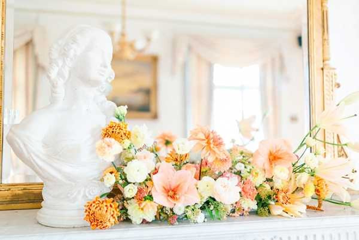 A close-up detail shot of a floral arrangement displayed on a white marble mantelpiece inside what appears to be a classic French-style interior with gold-framed mirrors and white paneled walls visible in the background. A white marble or plaster bust sculpture of a woman in 18th-century style is positioned to the left of the arrangement. The low, sprawling floral design features peach dahlias, blush amaryllis, ivory garden roses, orange chrysanthemums, cream ranunculus, and small yellow accent blooms with green foliage, creating a warm peach, apricot, and cream color palette. The overall decor styling is classic and formal, consistent with a chateau or period manor house setting.