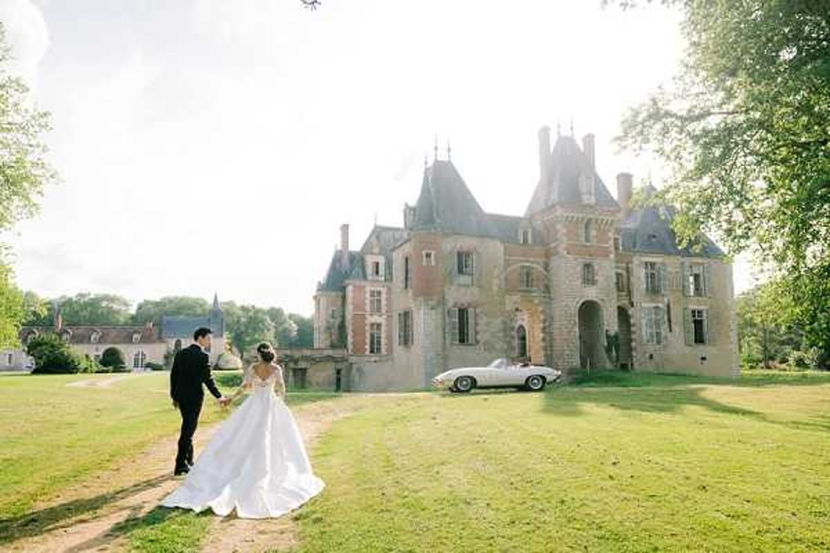 A bride and groom walk hand-in-hand across a wide lawn toward a French Renaissance-style chateau with stone and brick façade, pointed turrets, and arched entryway. The bride wears a long-sleeved white ballgown with a cathedral-length train, and the groom is dressed in a black tuxedo. A vintage cream-colored convertible sports car is parked in front of the chateau entrance, adding a classic styling element. The shot is a wide full-length portrait taken from behind the couple, emphasizing the grand scale of the chateau grounds and the sweep of the bridal gown. Potential venue feature image.