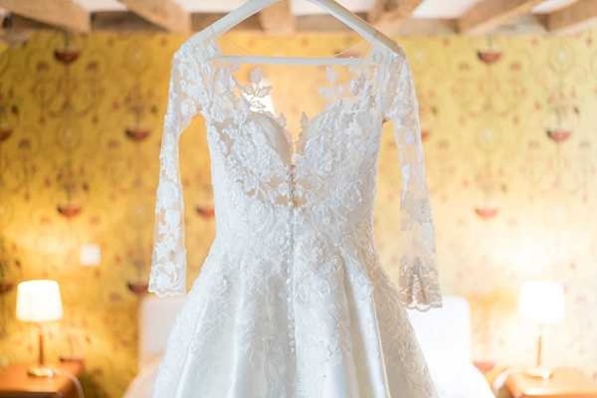 A close-up detail shot of an ivory lace wedding dress hanging on a white hanger against a yellow floral-patterned wallpaper. The gown features long illusion lace sleeves, a deep V-neckline with lace appliqué overlay, a fitted bodice with button detailing down the back, and a full A-line skirt. The room behind shows warm lamp lighting on bedside tables, suggesting a getting-ready setting inside a chateau or country house with rustic exposed ceiling beams visible at the top of the frame.