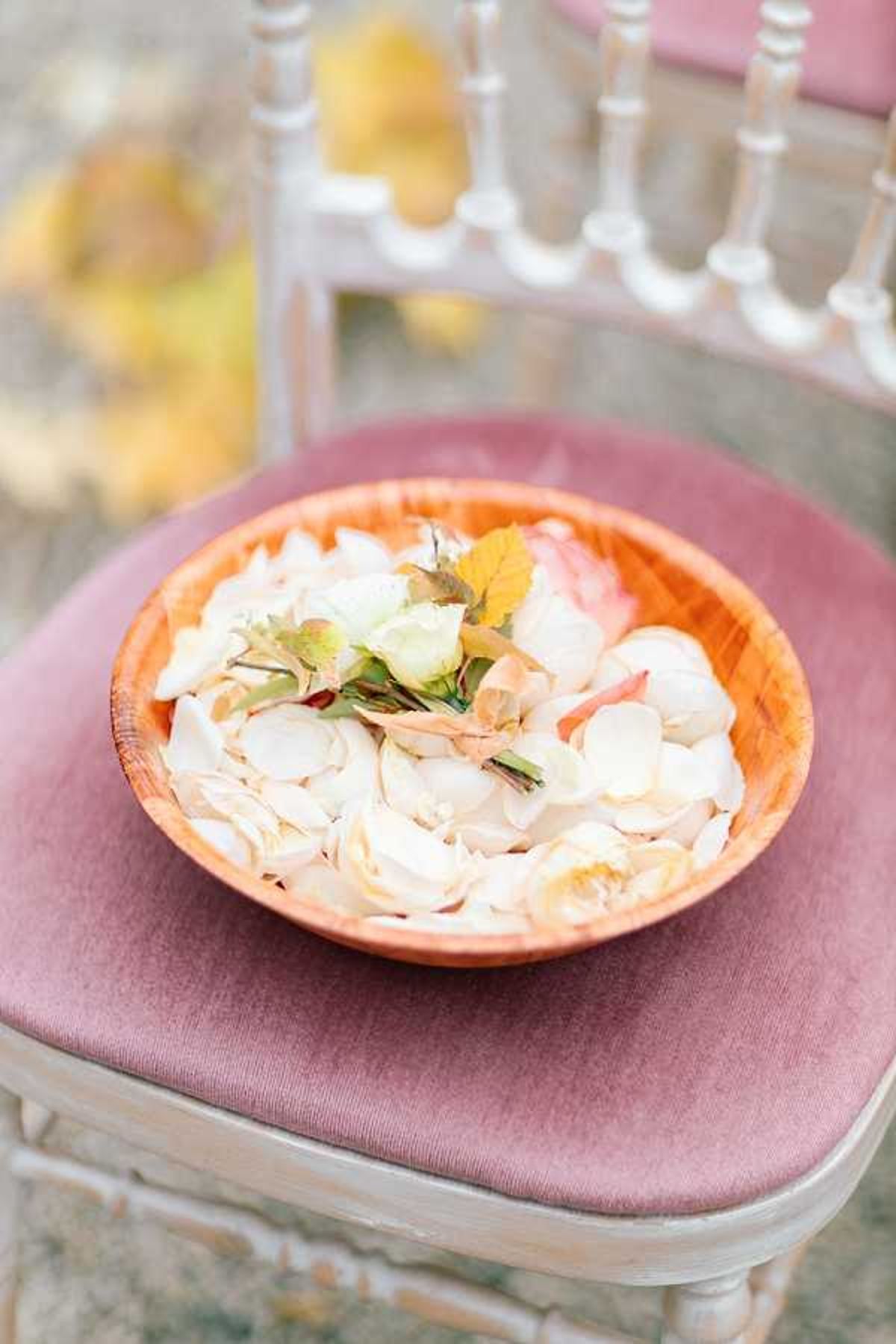A close-up detail shot of a wooden bowl filled with ivory and blush rose petals, including a few small whole flower heads with green foliage, placed on the dusty mauve velvet cushion of a whitewashed Chiavari chair. The setting appears to be an outdoor ceremony, with additional Chiavari chairs visible and softly blurred in the background. The petal toss styling coordinates with a romantic, classic palette of ivory, blush, and mauve. The composition is a tight portrait-orientation detail shot with shallow depth of field.