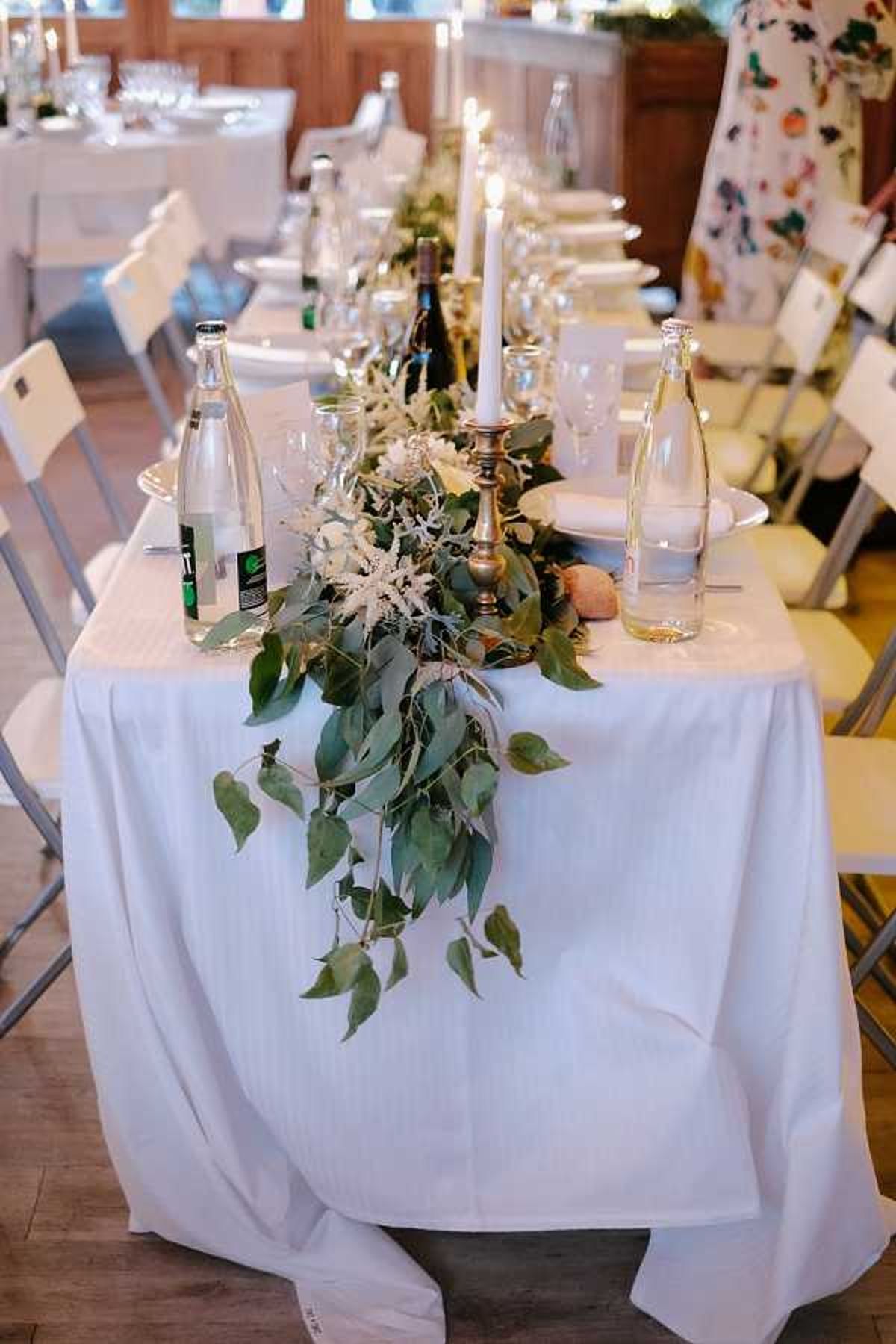 Close-up detail shot of a long reception dining table set up indoors in a wood-paneled room with warm lighting. The table is covered in a white linen tablecloth and features a lush greenery runner made of large-leafed branches and small white astilbe-style blooms cascading off the end of the table. Brass candlestick holders with tall white taper candles run along the center, accompanied by glass water bottles, wine bottles, crystal stemware, and white plates with folded napkins. White folding chairs line both sides of the table, with additional tables visible in the background. The overall decor palette is white and green with brass accents, leaning toward a natural, organic styling approach.
