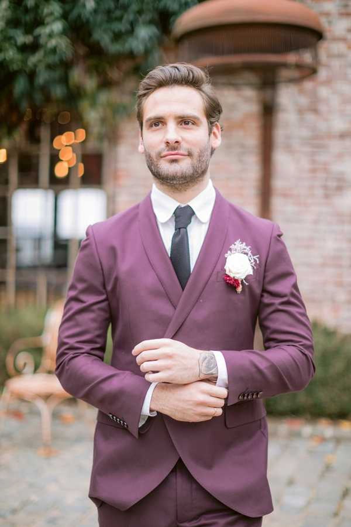 A groom portrait shot outdoors in a courtyard setting with brick architecture and warm bokeh lights visible in the background. The groom wears a fitted deep plum double-breasted suit with a white dress shirt and a dark charcoal knit tie, and his boutonniere features a cream garden rose with small burgundy floral accents. He is adjusting his cufflinks and has a visible tattoo on his hand, with a well-groomed beard and styled hair. The composition is a medium close-up portrait with a shallow depth of field that keeps the focus on the groom's face and attire.