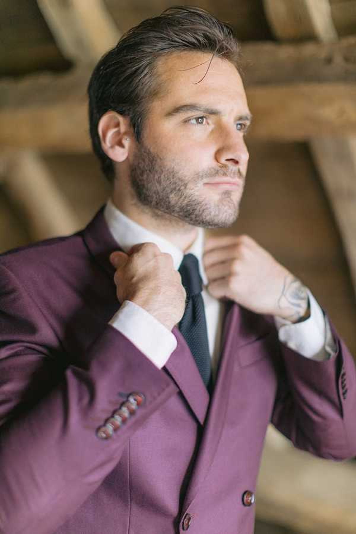A groom is photographed getting ready, adjusting his dark navy knit tie against a white dress shirt. He is wearing a deep burgundy double-breasted suit jacket with decorative multicolored boutonniere pins on the lapel pocket and matching burgundy buttons. The setting appears to be an indoor rustic space with exposed wooden ceiling beams visible in the background. The portrait is a close-up shot with a shallow depth of field, focusing on the groom from the chest up, with the groom's visible forearm tattoo adding a personal detail to the styling.