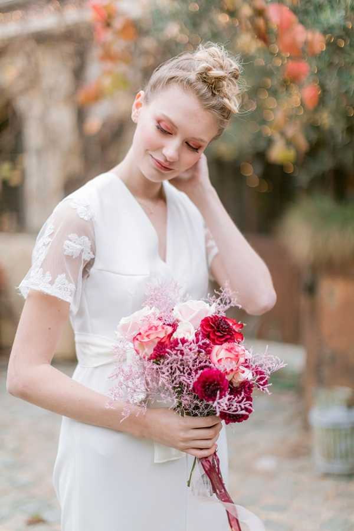 A bridal portrait of a single bride photographed outdoors in a courtyard setting with warm, blurred foliage and fairy lights visible in the background. The bride wears a fitted white crepe gown with a V-neckline, sheer flutter sleeves featuring white lace leaf appliqués, and a satin bow at the waist, with her blonde hair styled in a loose updo. She holds a compact bouquet composed of deep burgundy dahlias, blush and coral roses, and pink astilbe-style filler flowers, tied with a dark burgundy ribbon. The portrait is a close-up mid-shot with a shallow depth of field, capturing a romantic autumn color palette with rosy eye makeup complementing the bouquet's tones.