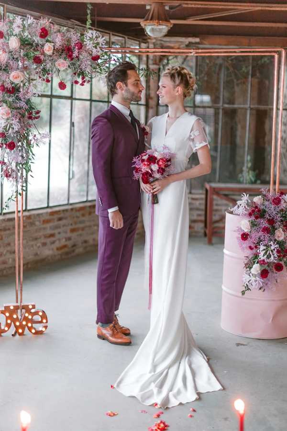 A couple stands together during what appears to be an indoor ceremony or styled shoot in an industrial-style venue with exposed brick walls, large metal-framed windows, and a concrete floor. The groom wears a deep plum/burgundy suit with brown oxford shoes, while the bride wears a fitted ivory satin gown with short sheer cap sleeves and a low back, her hair styled in an updo braid. She holds a bouquet of deep red dahlias, blush roses, and purple-pink wildflowers with long trailing ribbons. Behind them is a copper pipe arch decorated with blush roses, deep red blooms, and trailing greenery, and to the right stands a pink painted metal drum topped with a matching floral arrangement. A light-up marquee 'love' sign sits on the floor to the left, and lit candles with scattered rose petals are visible in the foreground. The overall decor palette combines copper, blush pink, and deep burgundy-red in a modern industrial style. Medium portrait shot.