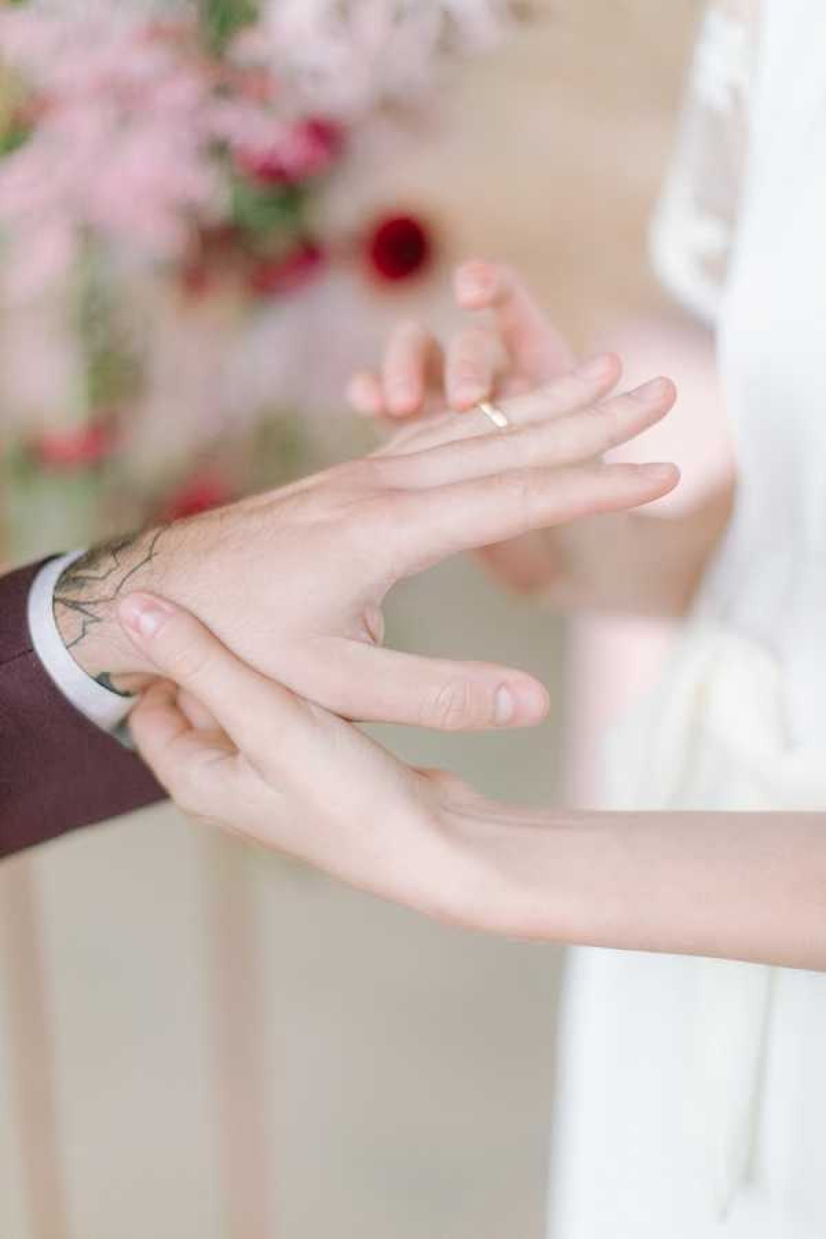 Close-up detail shot of a ring exchange moment during a wedding ceremony, showing the groom sliding a thin gold band onto the bride's finger. The groom's hand is visible at the left edge of the frame wearing a dark burgundy suit jacket with a white shirt cuff, and has a black tattoo on his wrist. Soft blurred floral arrangements in the background feature pink blooms and deep red flowers against a neutral, light backdrop. The bride is wearing a white dress visible at the upper right. The overall color palette is soft and muted with the burgundy suit and pink florals providing contrast.