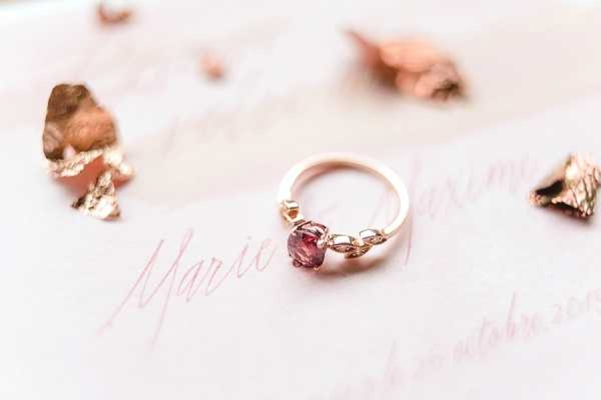 Close-up detail shot of a rose gold engagement ring featuring a round pink-to-mauve gemstone center stone with small diamond leaf-shaped accent settings on either side, resting on white wedding stationery with rose gold calligraphy script reading 'Marie' and 'Maxime' along with a date in October. Scattered around the ring are small dried rose petals gilded in rose gold, adding to the overall rose gold and blush pink color palette. The composition is a styled flat lay with a shallow depth of field, keeping the ring in sharp focus against the soft background of the stationery.