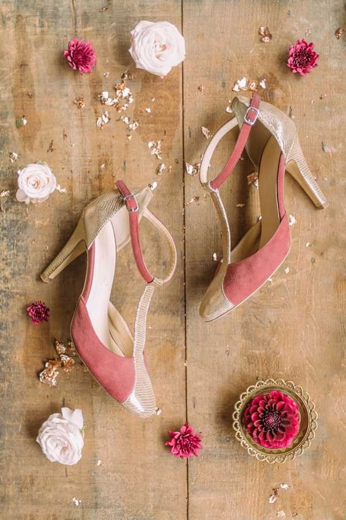 A flat-lay detail shot of a pair of bridal heels arranged on a rustic wooden surface. The shoes feature dusty rose suede toe caps with gold metallic snake-textured straps, ankle buckles, and gold heels. Scattered around the shoes are small magenta chrysanthemums, blush garden roses, and flecks of gold leaf confetti. A small ornate gold tray holding a single magenta chrysanthemum is placed in the lower right corner, tying the color palette together. The overall styling combines a dusty rose and gold color scheme with a romantic, slightly bohemian aesthetic.