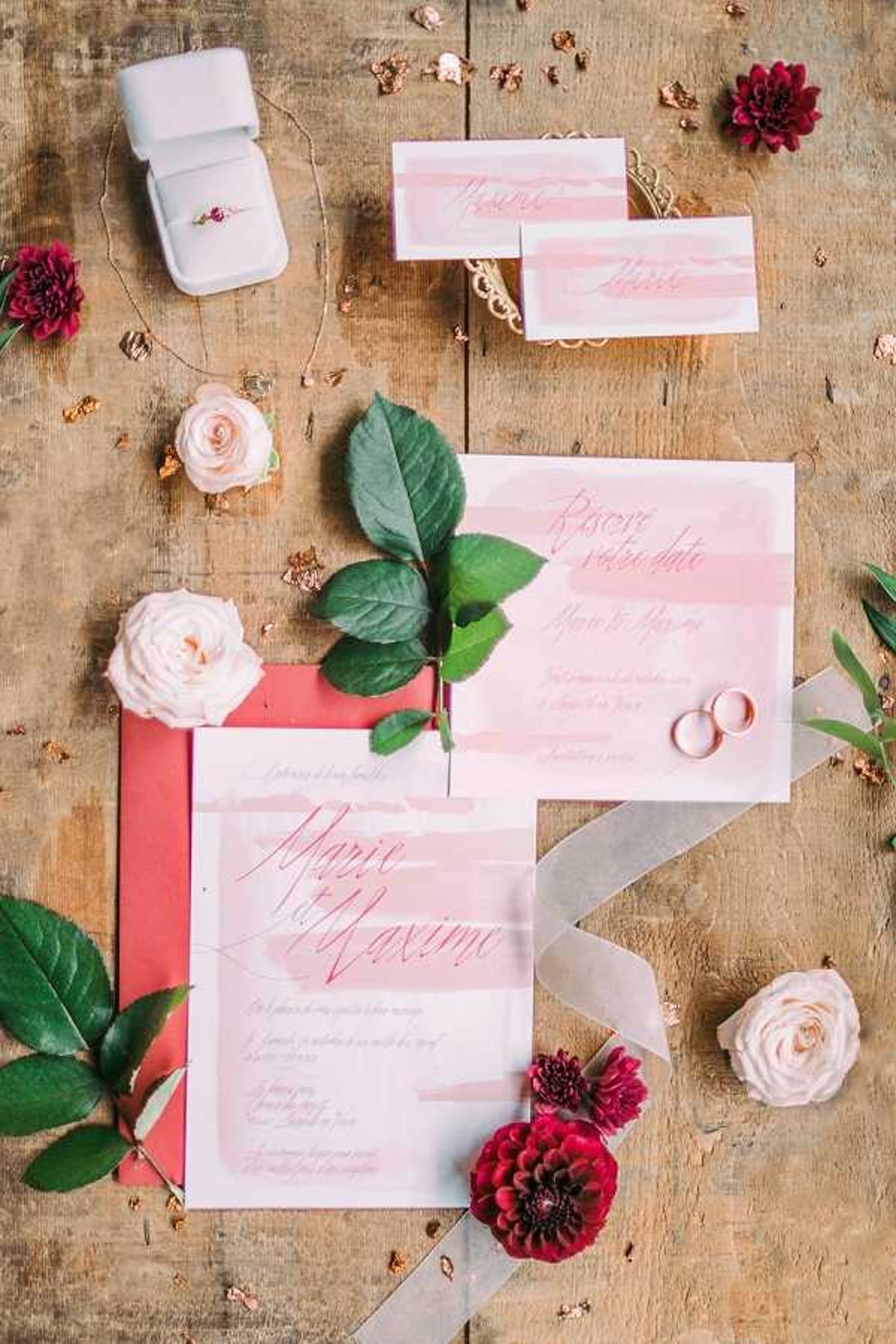 A flat lay detail shot of a French wedding stationery suite arranged on a weathered wooden surface. The suite includes a main invitation card and a save-the-date card for Marie and Maxime, both featuring blush pink watercolor stripe backgrounds with deep pink calligraphy script, along with a coral/red envelope and place cards in a matching style. Two gold wedding bands rest on one of the cards, and a ring box containing a ring with a red gemstone accent sits in the upper left corner. The arrangement is styled with cream garden roses, deep burgundy dahlias, green foliage sprigs, scattered gold leaf flakes, and a sheer gray ribbon. The overall color palette combines blush pink, deep burgundy, coral red, and gold.