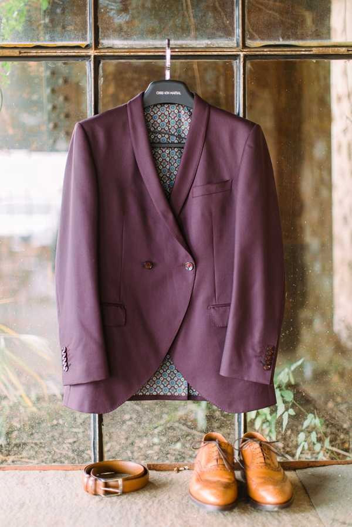 A groom's getting-ready detail flat lay featuring a deep burgundy double-breasted suit jacket by Chris Von Harsal, hung on a branded hanger against an aged industrial-style multi-pane window. The jacket has a shawl lapel, burgundy buttons, and a teal and rust geometric patterned lining visible at the interior. Below the jacket, a tan leather belt and a pair of tan leather wingtip brogue Oxford shoes are arranged on the floor. The overall styling has a modern, fashion-forward aesthetic with a rich jewel-tone color palette.