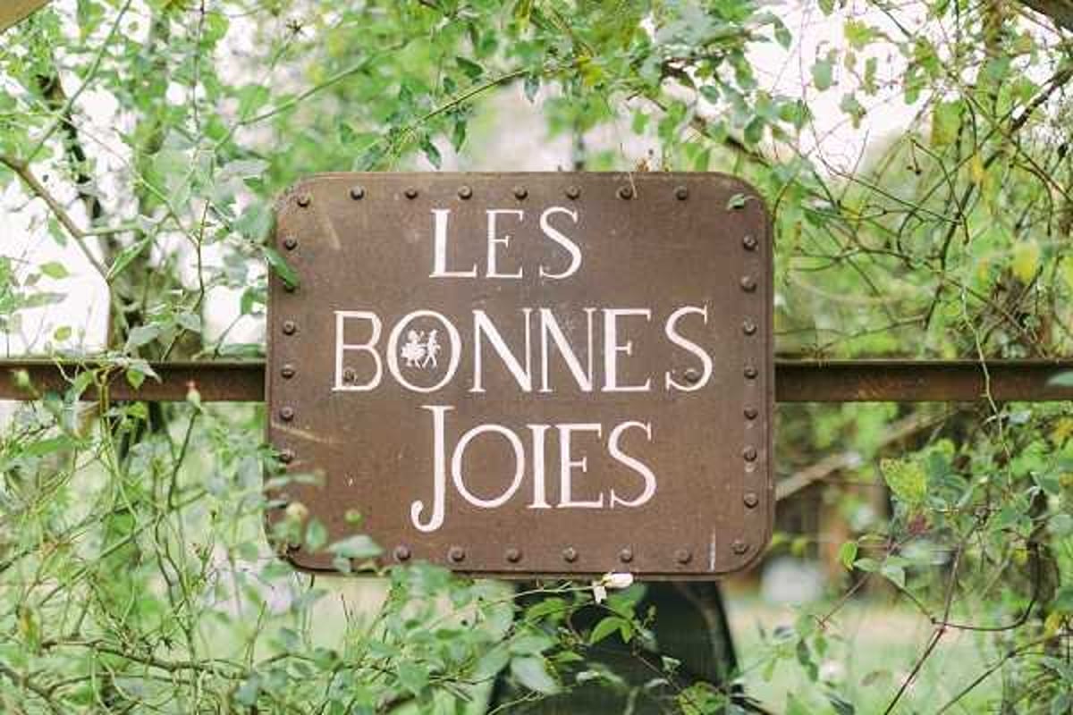 Close-up detail shot of a rectangular metal venue sign reading 'Les Bonnes Joies' in white lettering, mounted on a metal gate or fence and surrounded by green foliage. The sign has a dark brown, aged finish with riveted edges and features a small illustrated logo of two dancing figures. The composition is centered and tightly framed on the sign. Potential venue feature image.