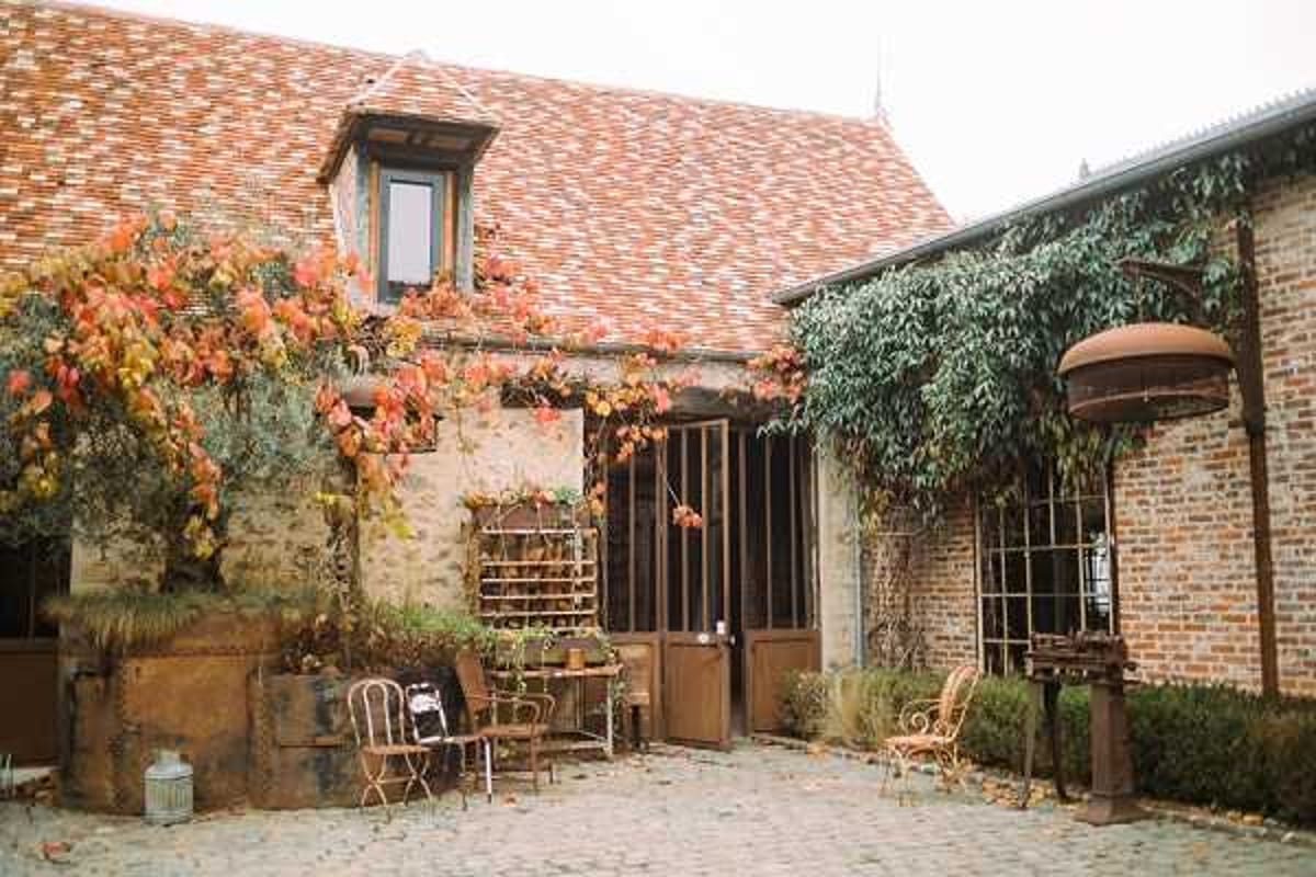 Wide shot of a rustic French farmhouse or domaine courtyard set up for a wedding, featuring a cobblestone forecourt with vintage wrought-iron bistro chairs and small decorative tables arranged outside wooden barn-style doors. The brick and stone building has a terracotta-tiled roof and is partially covered with climbing vines displaying red and orange autumn foliage. Decorative elements near the entrance include a wooden shelving display and a lantern placed on the ground, suggesting a cocktail hour or welcome area styled with a rustic, vintage theme. Potential venue feature image.