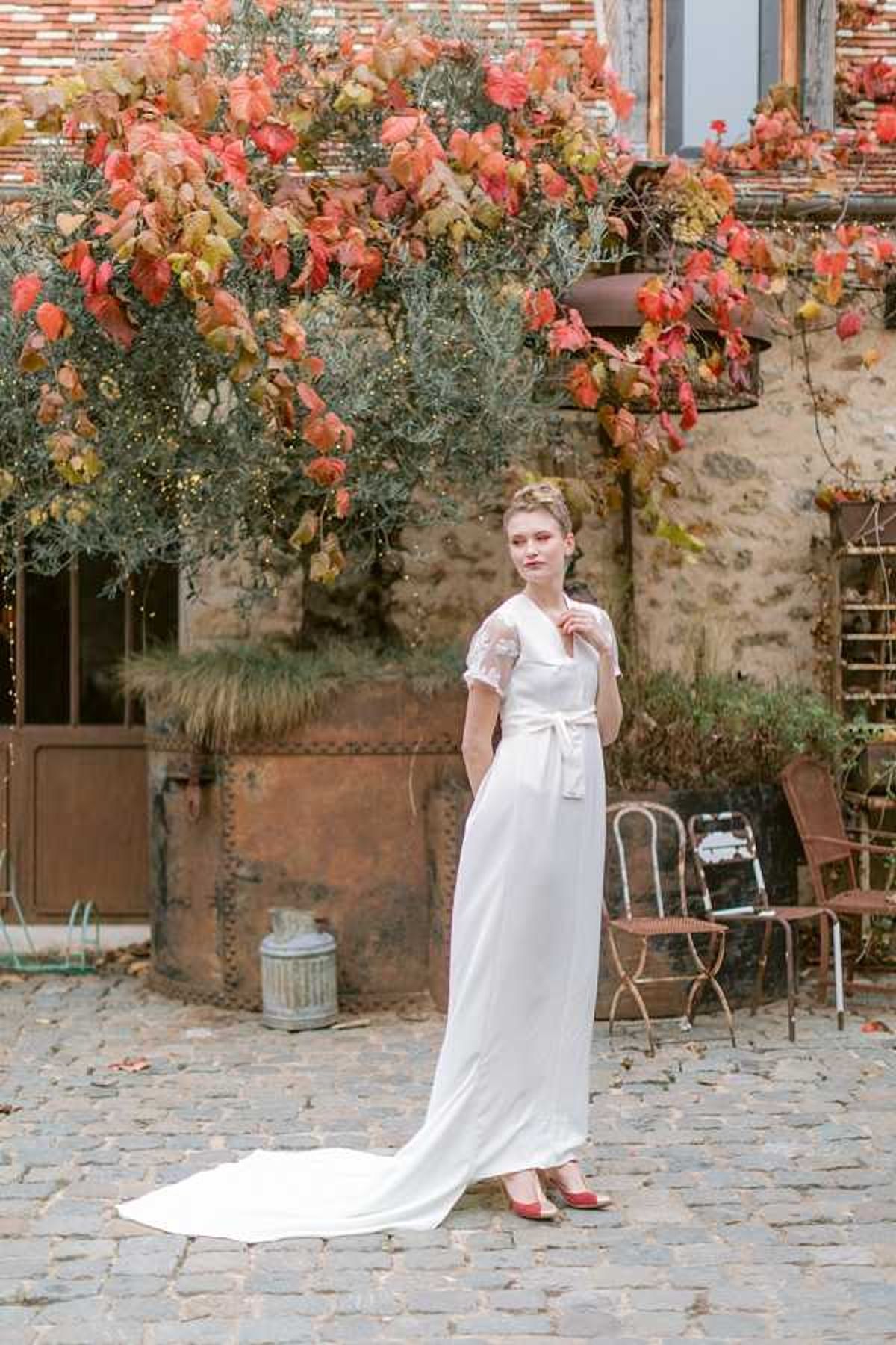A bridal portrait shot outdoors in a cobblestone courtyard of a rustic stone building, likely a French farmhouse or domaine. The bride stands alone wearing a sleek ivory crepe gown with short lace-embroidered sleeves, a white satin sash tied at the waist, and a flowing train, paired with red heeled shoes. Her hair is styled in an updo with a braided detail. The courtyard background features vintage-style metal bistro chairs, a galvanized watering can, and climbing vines with red and orange autumn foliage covering the building facade, giving the scene a warm autumnal palette. The composition is a full-length portrait with a slightly editorial feel, styled in a modern yet relaxed French aesthetic.