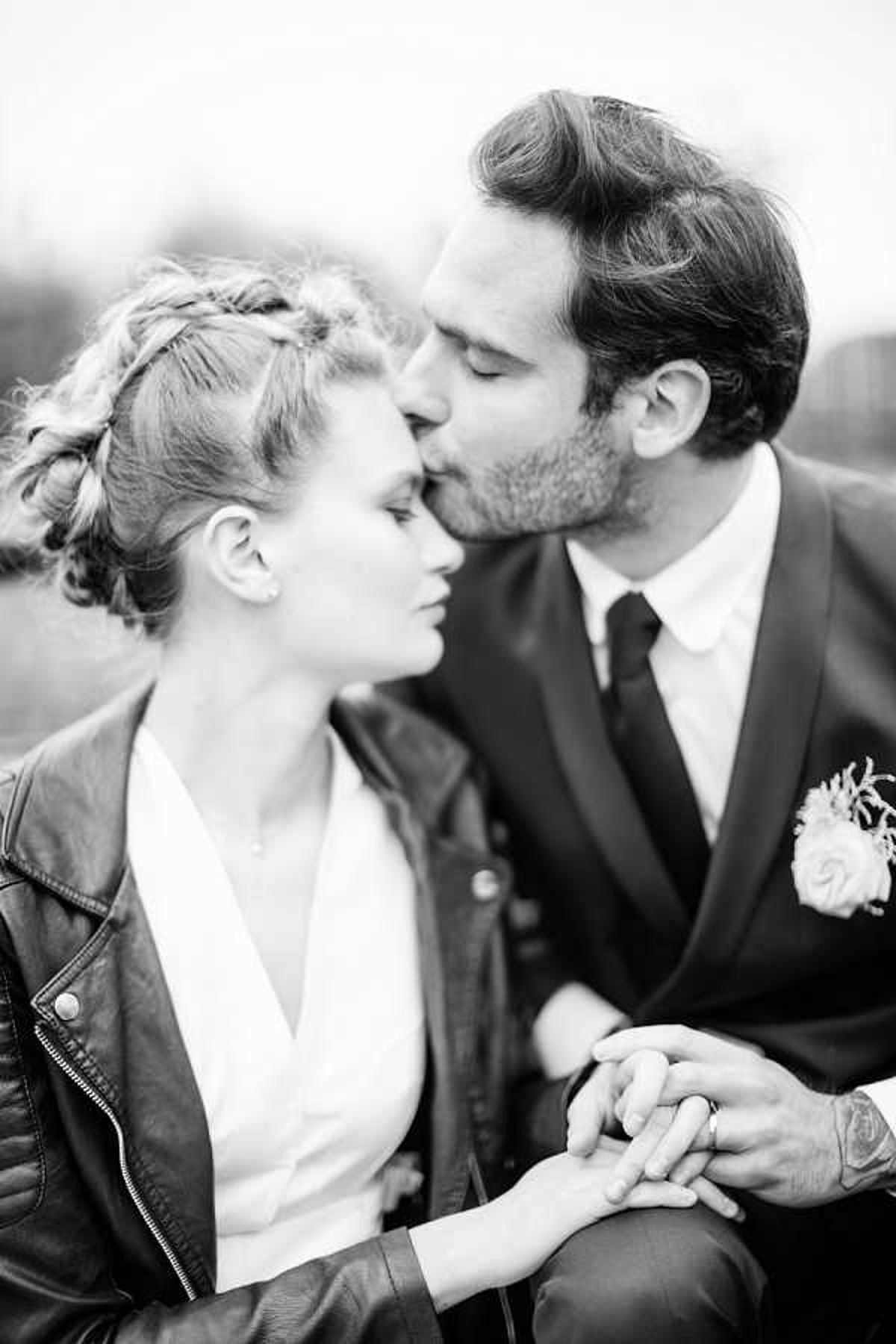 A black-and-white close-up portrait of a bride and groom sharing a kiss during what appears to be a couples portrait session outdoors, with a softly blurred background. The bride wears a white V-neck top or dress paired with a leather moto jacket and has her hair styled in a braided updo; the groom wears a dark suit with a dark tie and a boutonnière featuring a small rose and textured foliage. The couple holds hands in their laps, with a tattooed wrist visible on the groom. The image has soft, high-key tones with gentle contrast, giving it a light, airy feel consistent with a modern, slightly edgy styling aesthetic.