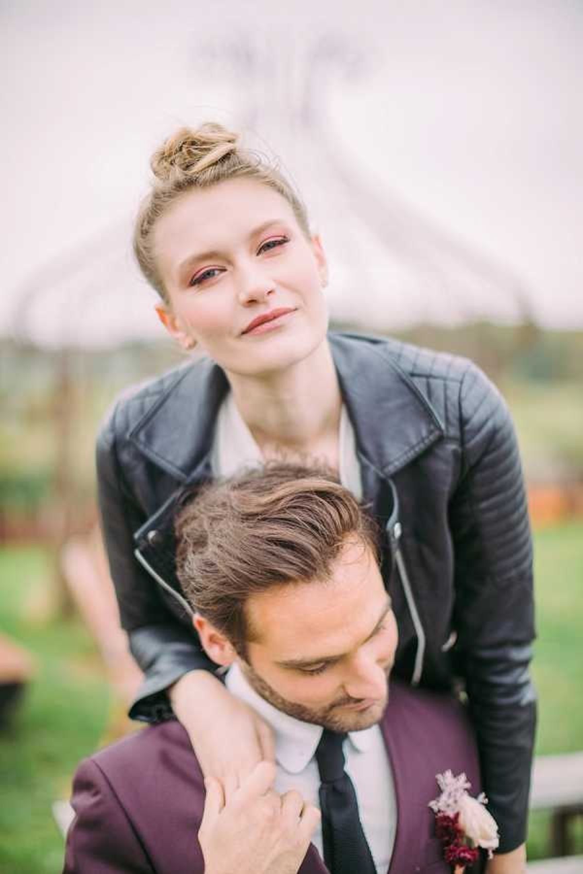 A couple portrait shot outdoors, with the bride leaning over the groom's shoulder from behind in a playful pose. The bride wears a black leather moto jacket over her wedding dress, her blonde hair styled in a top knot, and bold reddish-pink eye makeup. The groom wears a deep plum/burgundy suit with a white dress shirt, black tie, and a boutonnière featuring blush pink roses and deep burgundy florals. The styling is distinctly edgy and modern, contrasting the classic bridal context. The composition is a close-up portrait with shallow depth of field, blurring the outdoor background which appears to include a tent or tipi structure.