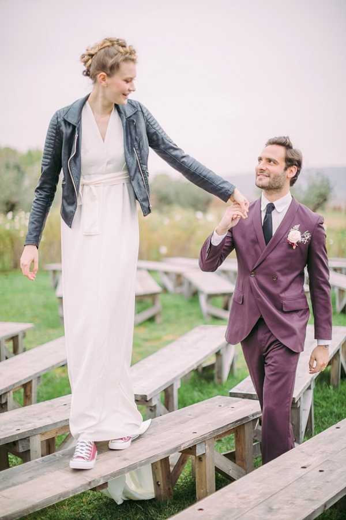 A couple portrait shot outdoors in what appears to be a ceremony seating area with rows of rustic wooden benches set on grass. The bride stands on one of the benches, balancing while holding the groom's hand; she wears a simple ivory wrap-style floor-length dress with a sash belt, a dark navy leather moto jacket, a braided updo, and burgundy Converse sneakers. The groom wears a double-breasted plum/mauve suit with a black tie and a blush pink boutonniere featuring small florals. The overall styling is modern and casual with eclectic, non-traditional elements. The shot is a full-length portrait with a slightly overcast outdoor backdrop.