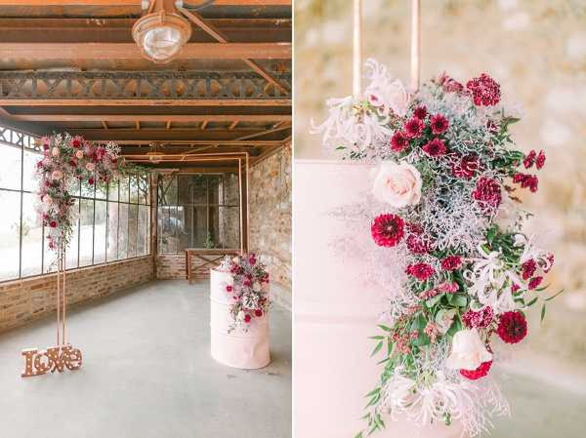 A two-panel detail shot showcasing wedding decor styled in an industrial indoor venue with exposed wooden ceiling beams, metal framework, and stone walls. The left panel shows a copper-toned metal arch decorated with cascading burgundy dahlias, blush roses, and trailing greenery, positioned beside a blush pink single-tier wedding cake on a matching pink cake stand; a wooden 'LOVE' sign in a copper finish sits on the concrete floor at the base of the arch. The right panel is a close-up of the wedding cake, which is a soft blush pink naked or semi-frosted cake adorned with a dramatic floral arrangement featuring deep burgundy dahlias, blush roses, white feathery thistle or similar textural blooms, and eucalyptus greenery cascading down one side. The overall decor palette combines copper metallics, blush pink, and deep burgundy/wine tones in a modern industrial-romantic style.