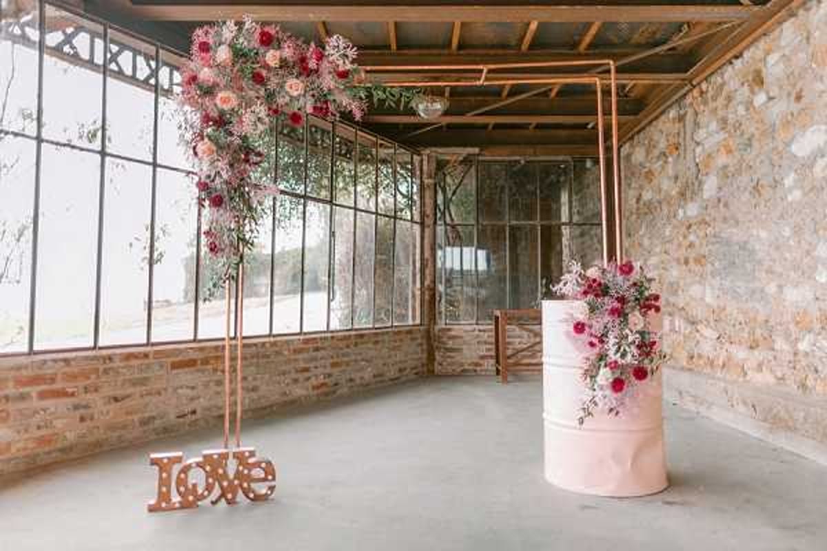 A styled wedding decor setup photographed in a wide shot inside an industrial-style venue with exposed stone walls, large metal-framed windows, and a concrete floor. A copper pipe arch stands on the left, topped with an asymmetric floral arrangement of blush pink, hot pink, and deep red blooms including roses and dahlias with trailing greenery, and a light-up wooden 'love' sign at its base. To the right, a blush pink cylindrical pedestal holds a coordinating floral arrangement in the same red and pink color palette. The overall styling theme is industrial-romantic, combining raw architectural elements with warm copper metalwork and a bold fuchsia and blush floral palette. Potential venue feature image.