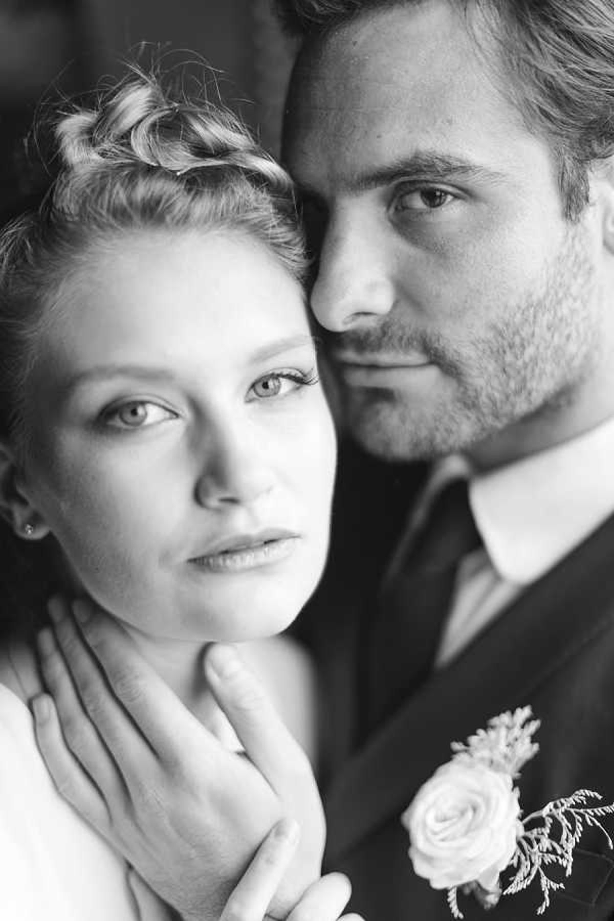 A black-and-white close-up portrait of a bride and groom posed together, both looking directly at the camera with a composed, serious expression. The bride has her hair styled in an updo with braided detail and wears small stud earrings, while the groom is dressed in a dark suit jacket with a tie and a boutonniere featuring a small rose and delicate foliage with fine textured greenery. The groom's hand rests gently against the bride's neck and shoulder. The image uses high contrast with bright highlights on the faces and soft, dark tones in the background, giving it a editorial, classic feel. The tight crop focuses entirely on the couple's faces and upper torsos.