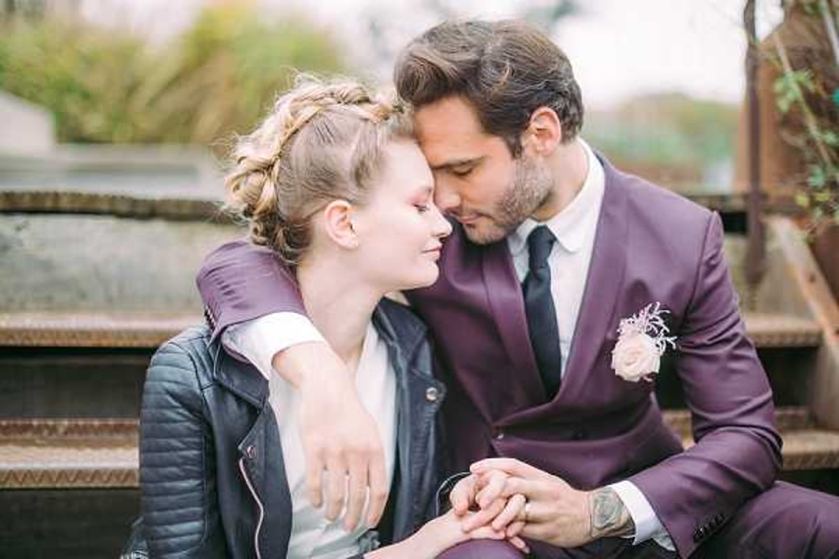 A close-up couple portrait of a bride and groom seated outdoors on what appears to be a metal staircase or industrial structure. The groom wears a deep plum/burgundy suit with a white dress shirt, black tie, and a blush pink boutonniere, while the bride wears a white dress layered under a black quilted leather jacket. The bride has her hair styled in a loose updo with soft face-framing pieces. The groom leans in close to the bride's cheek as she smiles with eyes closed, with his arm around her and their hands intertwined. The styling combines a classic bridal look with an edgy, modern aesthetic through the leather jacket pairing.