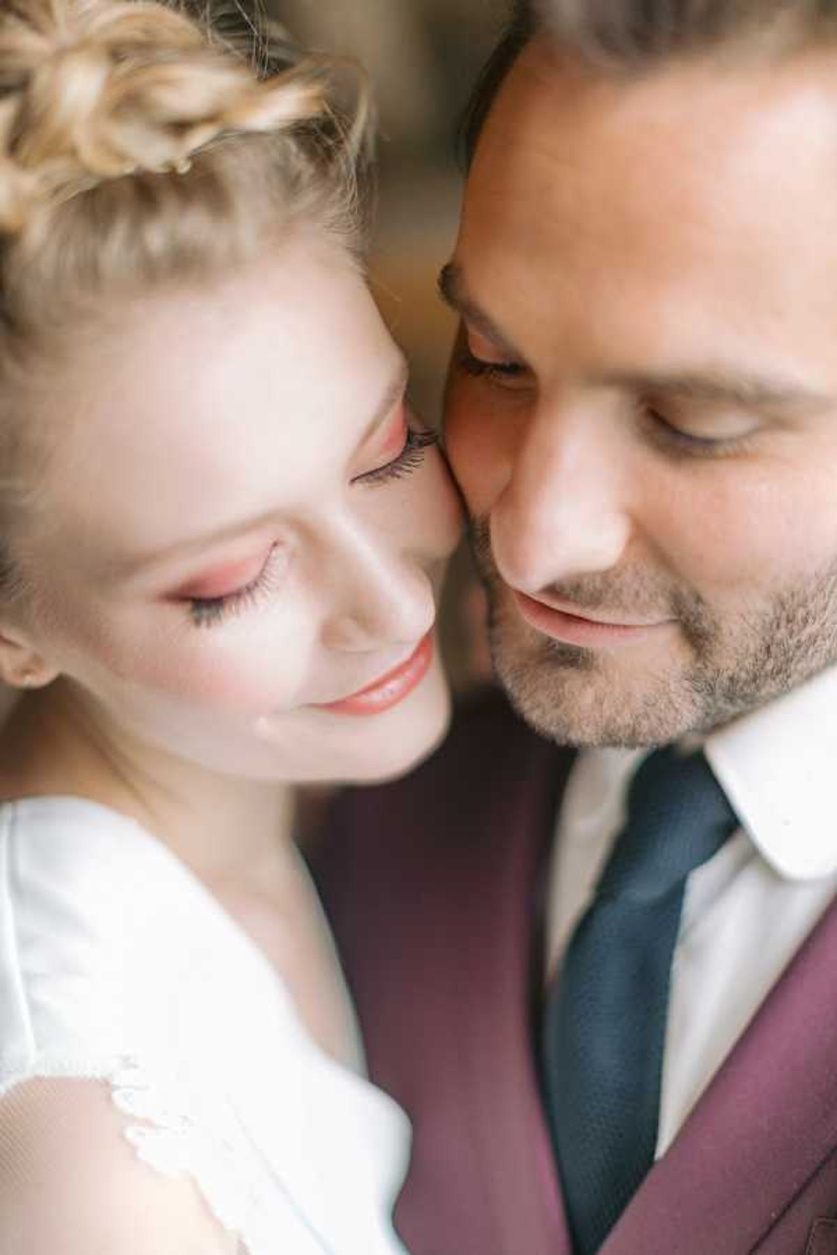 A close-up portrait of a bride and groom with their faces pressed together, both with eyes closed or downcast and smiling softly. The bride wears a white lace-detailed dress and has her blonde hair styled in a braided updo, with rosy pink eye makeup and a coral-tinted lip. The groom wears a burgundy suit jacket with a white dress shirt and a navy tie. The composition is tightly cropped to their faces and upper shoulders, with a shallow depth of field that softly blurs the background.