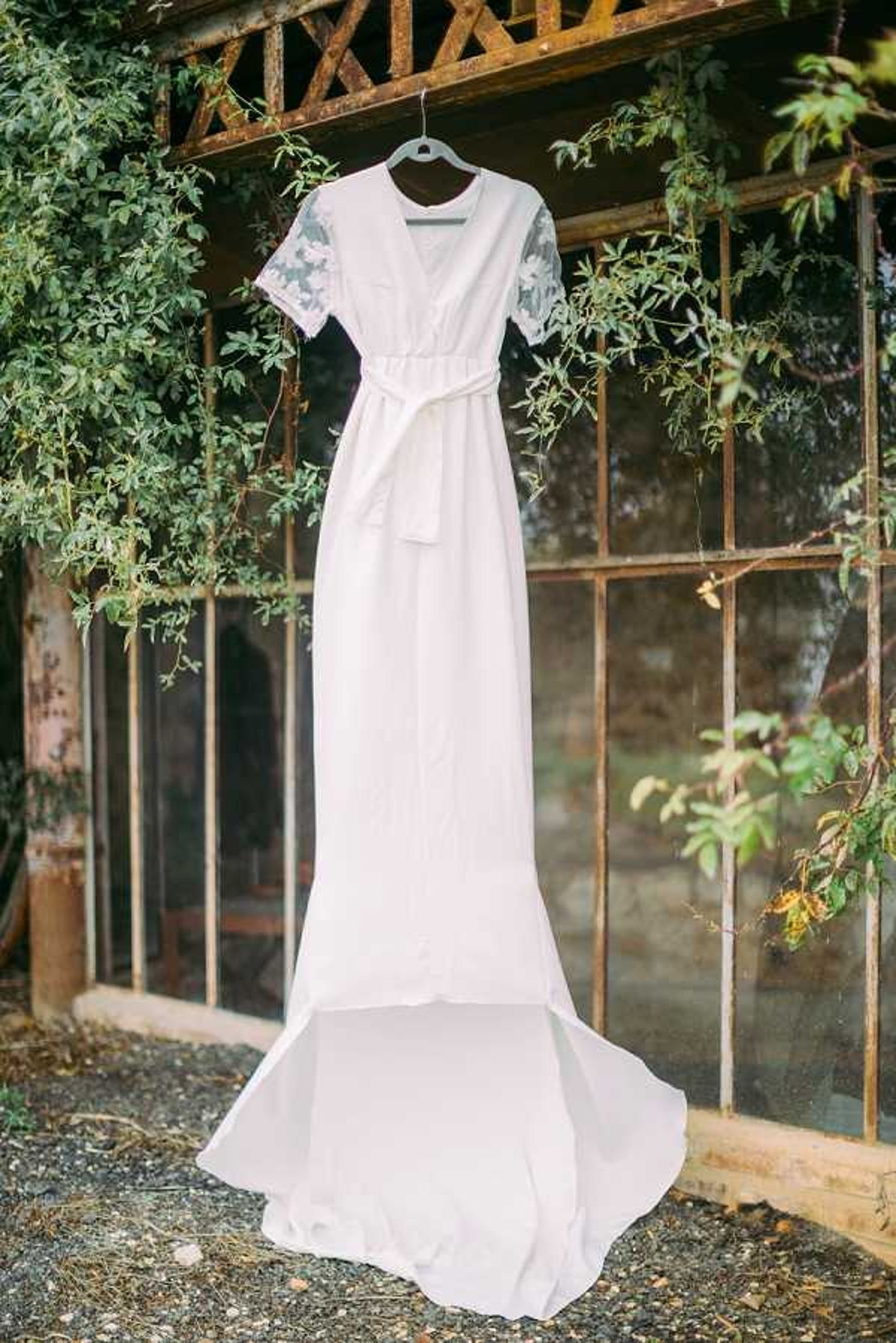A detail shot of a white bridal gown hanging on a hanger against the exterior of a rustic iron-framed greenhouse or orangerie structure with large glass panes. The dress features short lace sleeves with floral appliqué detailing, a V-neckline, a fitted silhouette with a self-tie satin belt at the waist, and a modest train pooling on the ground. The style is clean and minimalist with a boho influence. The iron framework of the structure is aged and patinated, and climbing vines frame the composition on either side.