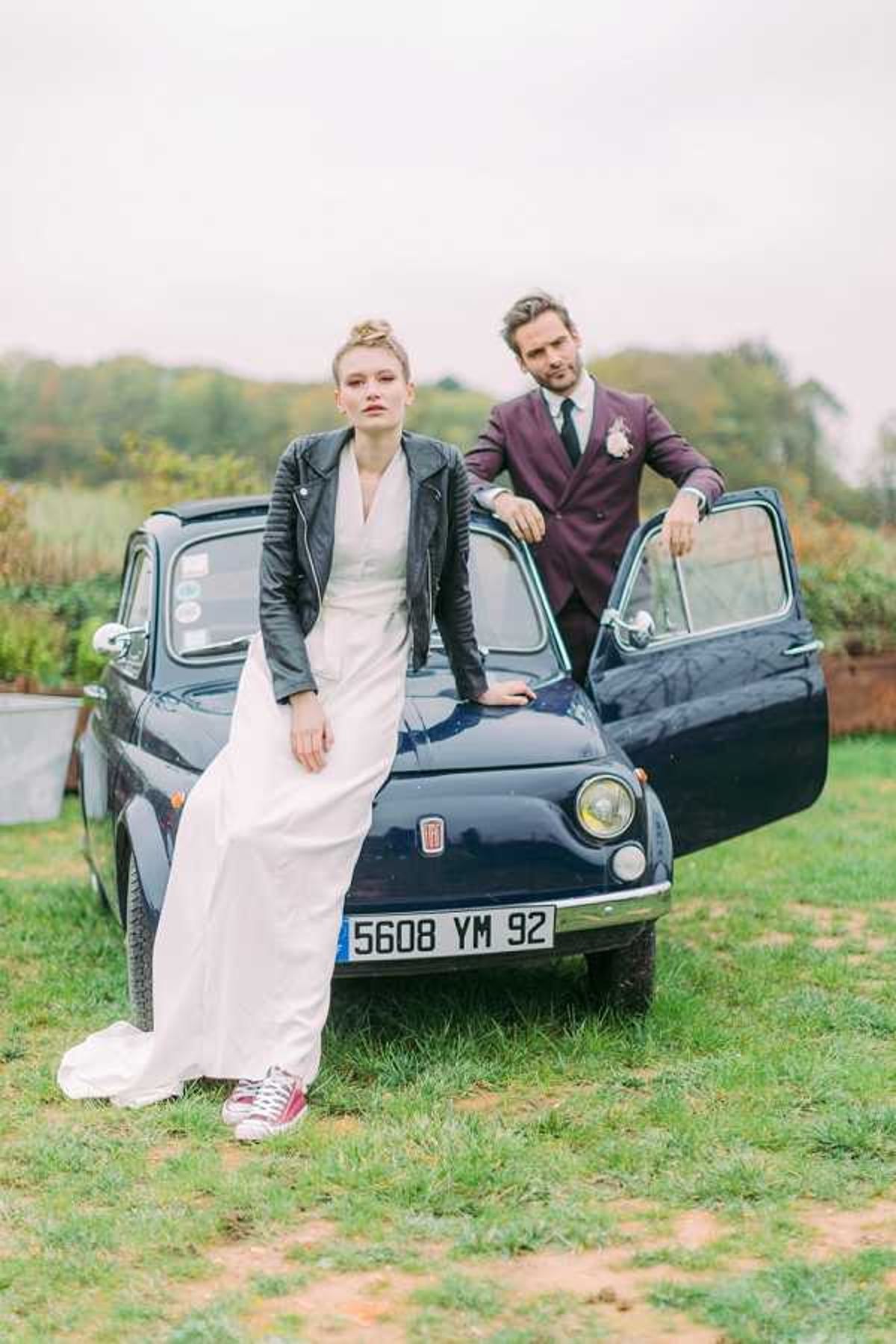 A couple poses outdoors in front of a vintage navy blue Fiat 500 with a French license plate, parked on grass. The bride wears a white column-style wedding dress paired with a black leather jacket and burgundy red Converse sneakers, with her hair styled in an updo; the groom stands behind the car with the door open, wearing a deep plum double-breasted suit with a dark tie and a small floral boutonniere. The styling has a distinctly edgy, modern aesthetic, blending bridal wear with streetwear elements. This is a full-length couple portrait shot in an outdoor setting with open countryside visible in the background.