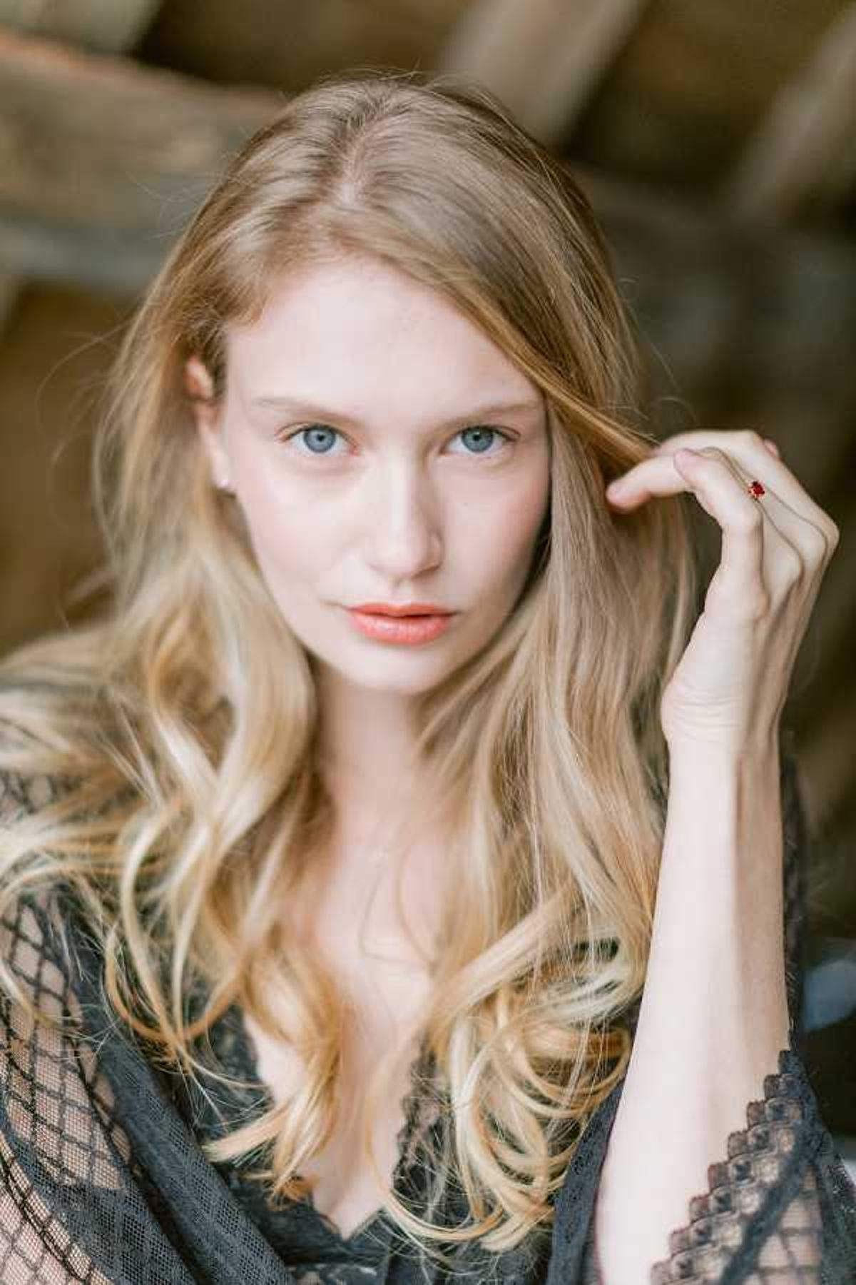 Close-up portrait of a woman with long, loosely waved blonde hair and blue eyes, wearing a black lace top or robe. She is touching her hair with one hand, on which a small red gemstone ring is visible. Her makeup is minimal with a peachy-coral lip. The background shows blurred wooden beams, suggesting a barn or rustic indoor setting. The shot is a shallow depth-of-field portrait with soft, natural light, likely from a getting-ready or bridal boudoir session.