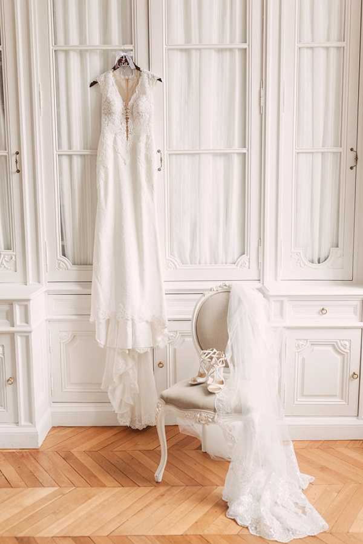 A bridal flat-lay style getting-ready shot set in an ornate white-painted room with floor-to-ceiling mirrored armoires and herringbone parquet flooring. An ivory lace wedding dress with a deep V-neckline, lace appliqué bodice, and tiered lace hem is hung from the top of the wardrobe. A white lace-edged veil is draped over a French-style carved white chair with a grey upholstered seat, and a pair of white strappy lace-up bridal heels rests on the chair seat. The overall palette is ivory and white against the warm wood flooring, with a classic Parisian interior aesthetic. Wide portrait-orientation shot capturing the full length of the dress and the room setting.