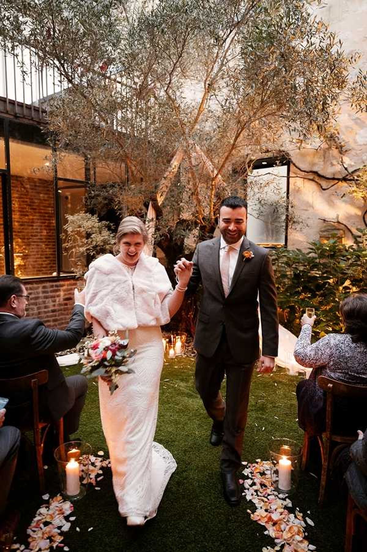 The bride and groom walk back up the aisle together following their ceremony in an indoor-outdoor courtyard venue with exposed brick walls, large steel-framed windows, and mature olive trees. The bride wears a fitted ivory lace gown paired with a white faux-fur stole and carries a bouquet of coral, burgundy, and blush florals with eucalyptus; the groom wears a charcoal suit with a pale pink tie and an orange buttonhole. The aisle is lined with pillar candles in glass hurricane holders and scattered peach and coral rose petals, creating a warm, candlelit atmosphere. Guests seated on wooden chairs flank the aisle, and the overall styling has an intimate, urban-chic feel; the image is a medium portrait shot taken from the front as the couple walks toward the camera.