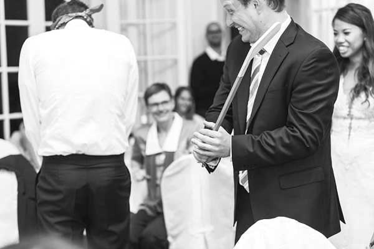A black-and-white candid reception moment captured in a mid-range portrait shot, showing a man in a dark suit and striped tie laughing and holding what appears to be a stick or cane, possibly during a game or comedic moment. A bride in a white dress is visible and smiling on the right edge of the frame, while seated guests laugh and react in the background. The setting appears to be an indoor reception room with large windows. The image has soft contrast with bright highlights, conveying a lively, joyful atmosphere among the group.