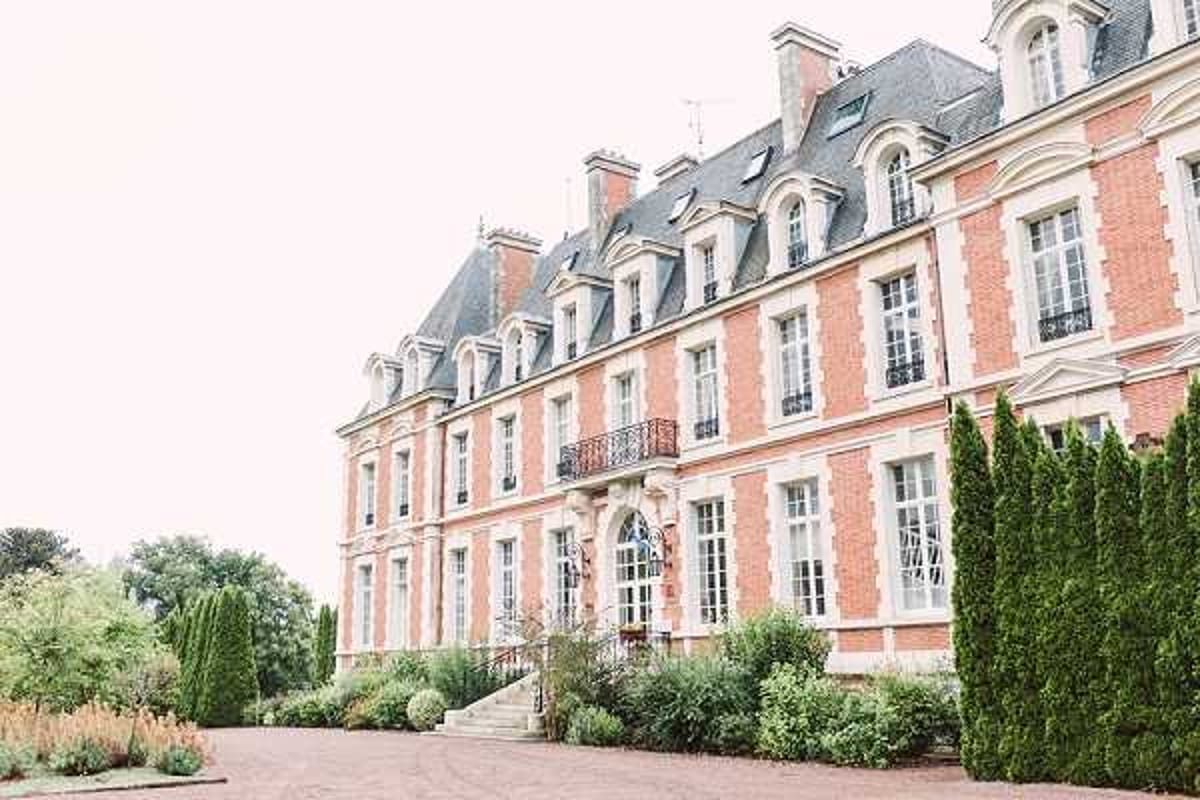 Wide exterior shot of a large French chateau featuring red brick facade with white stone trim, mansard roof with dormer windows, and ornate wrought-iron balconies. The building is three stories tall with symmetrical window arrangements and a central entrance framed by decorative stonework and steps. The foreground shows a gravel courtyard with manicured hedging and ornamental plantings. Potential venue feature image.