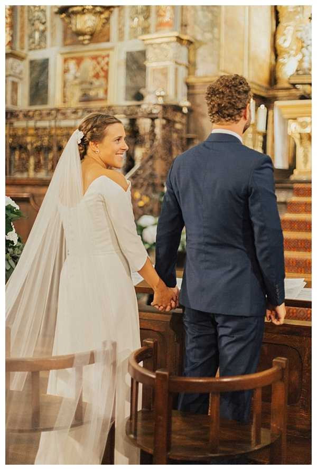 A couple stands at the altar during an indoor religious ceremony inside an ornate church with gilded baroque decor and painted panels visible in the background. The bride turns slightly toward the camera with a smile while holding the groom's hand; she wears a long-sleeved ivory gown with a low back, a cathedral-length veil, and a small white floral hair accessory. The groom, seen from behind, wears a navy blue suit. The composition is a medium shot taken from slightly behind and to the side of the couple, emphasizing their joined hands and the richly decorated church interior.