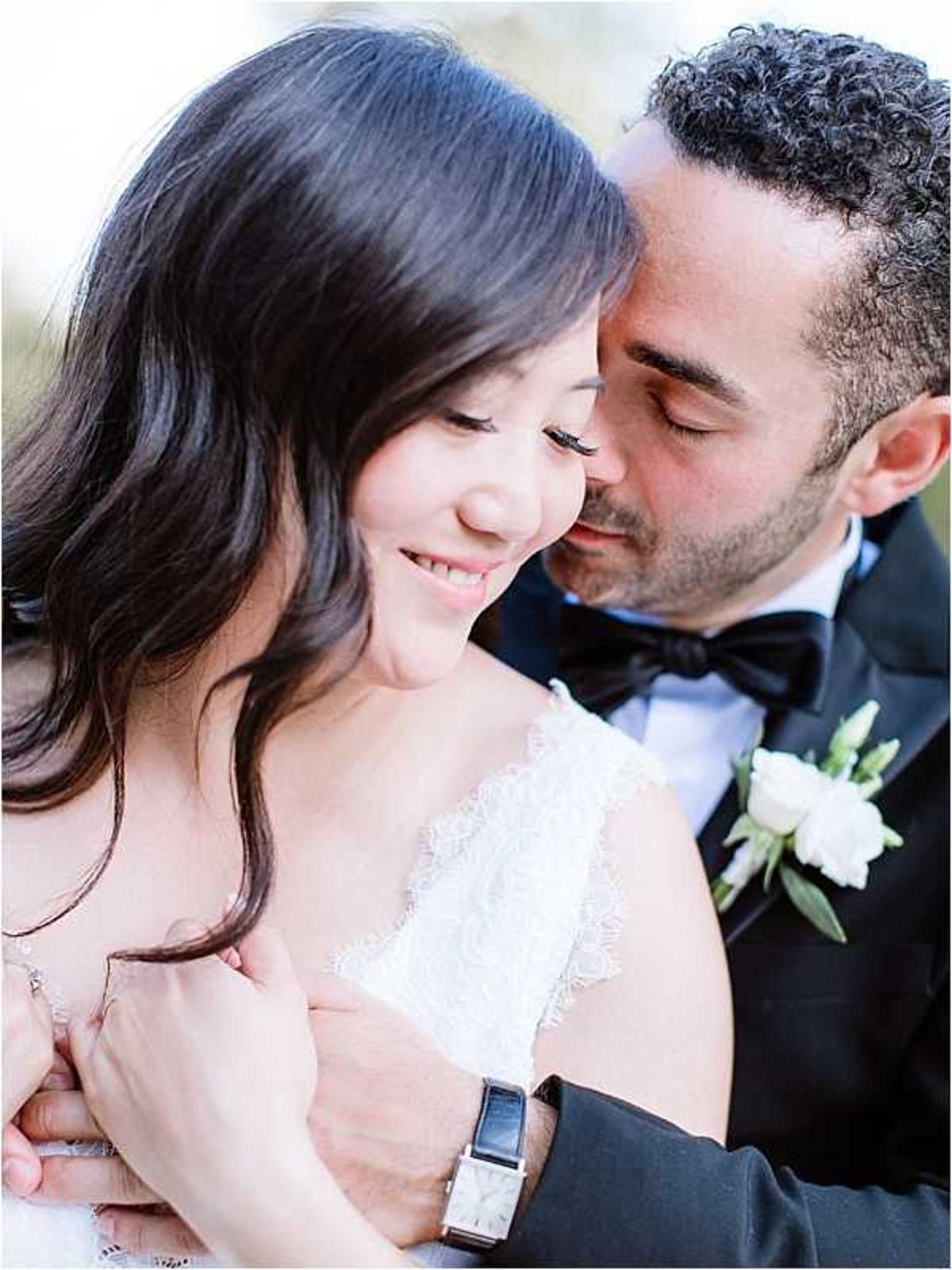 A close-up couples portrait showing the bride and groom in an intimate pose, with the groom leaning in close to the bride's cheek while she smiles softly with eyes downcast. The bride wears a white lace strapless or spaghetti-strap gown with scalloped lace detailing and has long dark hair worn loose. The groom is dressed in a black tuxedo with a black bow tie and wears a white rose boutonniere with small greenery accents; a rectangular-faced watch is visible on his wrist. The background is softly blurred with a light, airy tone, keeping the focus entirely on the couple. The overall styling is classic and refined, with a clean white and black color palette.