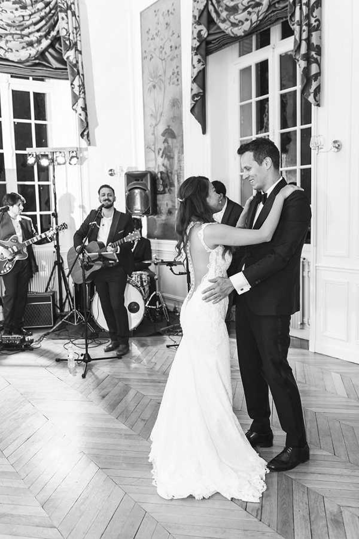 Black-and-white image of a couple sharing their first dance in an indoor ballroom with a classic parquet herringbone wood floor. The bride wears a form-fitting lace gown with an open back, and the groom is in a dark tuxedo with a bow tie and white French-cuff shirt; both are smiling and facing each other. In the background, a live band of at least three musicians performs, with two guitarists and a drummer visible, set up against a wall featuring tall white French doors and ornate patterned drapery with a decorative valance. The image is a medium-wide shot with strong contrast between the bright room and darker background tones.