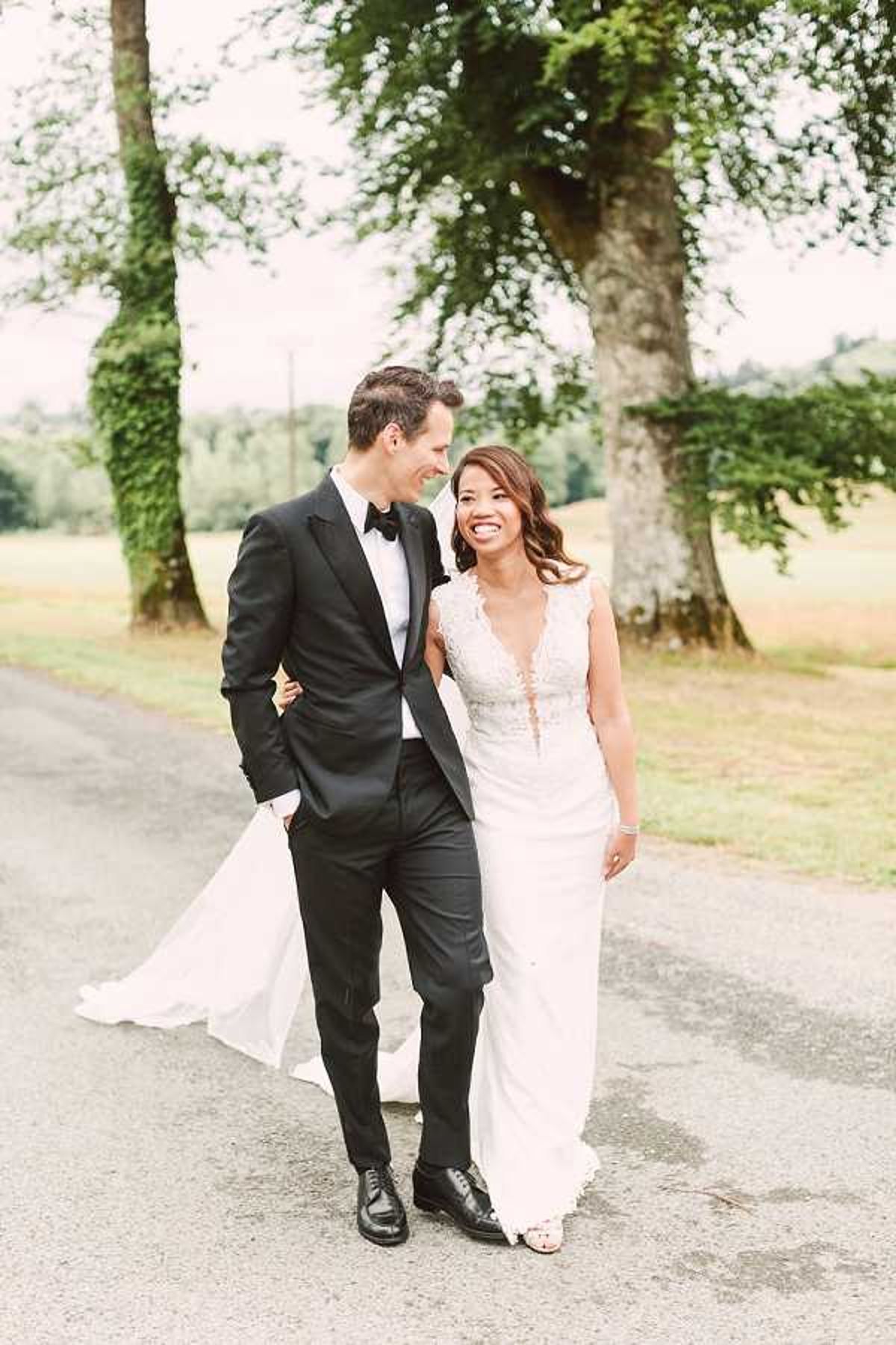 A couple portrait taken outdoors on a paved path, with the bride and groom walking together and looking at each other. The groom wears a classic black tuxedo with a black bow tie and black dress shoes, while the bride wears a sleeveless ivory gown with a deep V-neckline, lace bodice detailing, and a flowing train that the groom lightly holds. The setting appears to be the grounds of a French country estate or chateau, with a wide tree-lined driveway visible in the background. The overall styling is classic and formal, and the shot is a full-length portrait with a slightly soft, bright exposure.