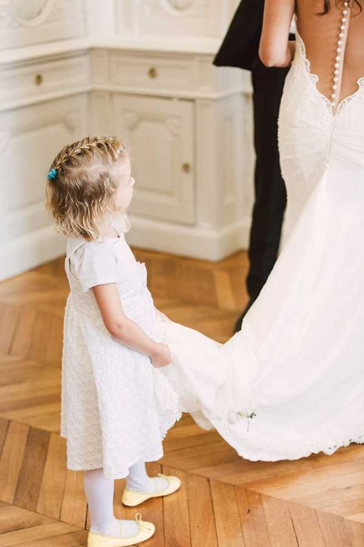 A candid portrait shot taken indoors showing a young flower girl, approximately toddler age, holding the train of a bride's white wedding gown. The flower girl wears a white eyelet broderie anglaise dress with short sleeves, white tights, and yellow flat shoes, with her blonde hair styled in a small braid accessorized with a blue clip. The bride wears a fitted white gown with a low back featuring a row of small pearl buttons and lace detailing along the neckline. The setting appears to be a classic French interior with white painted paneled cabinetry and warm-toned herringbone parquet flooring, consistent with a chateau or manor house getting-ready room. A figure in a dark suit is partially visible in the background.