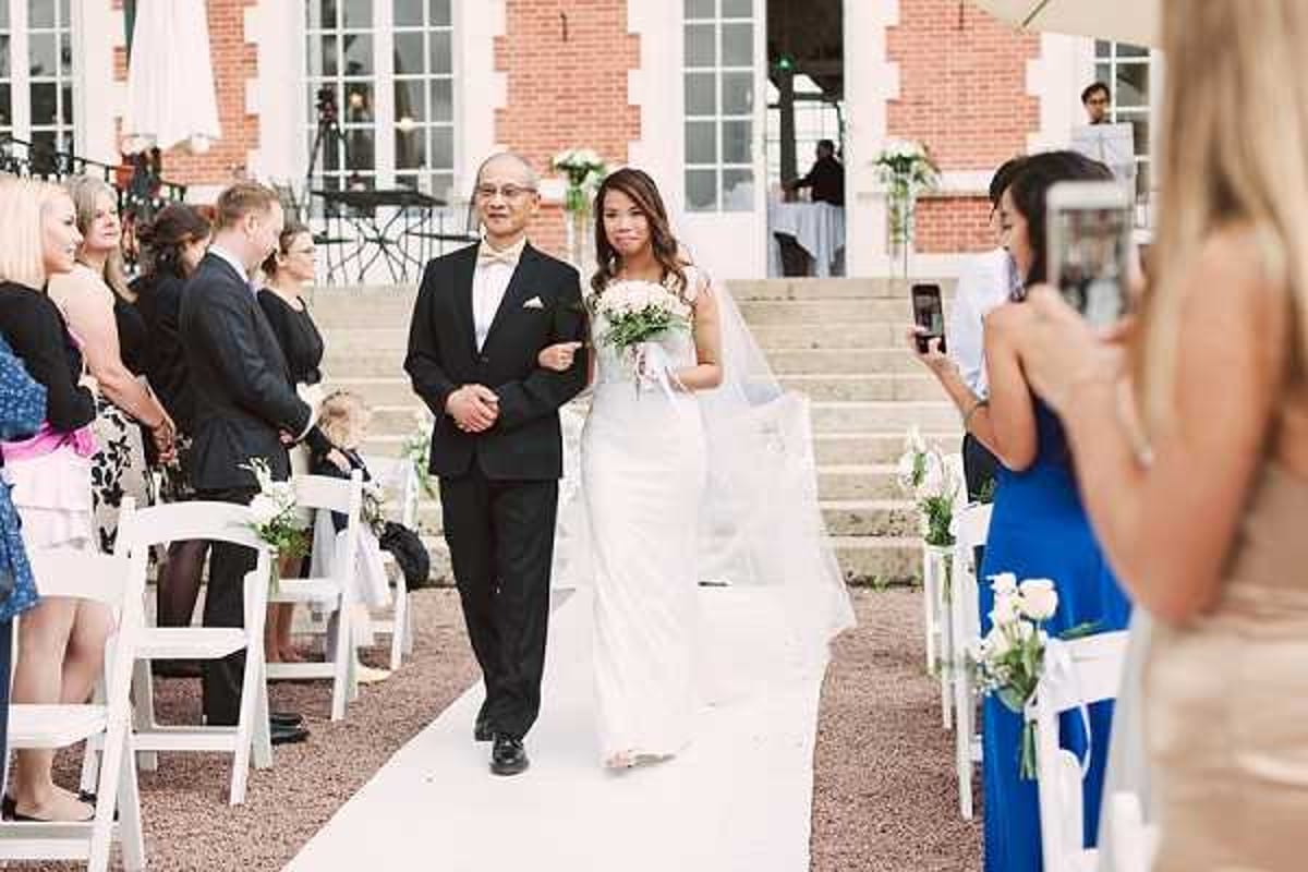 An outdoor ceremony processional showing a bride walking down the aisle escorted by an older man, likely her father, who is wearing a black tuxedo with a white pocket square. The bride is dressed in a slim-fit ivory gown with a long veil and carries a white bouquet. The aisle is lined with white folding chairs filled with seated guests, and white floral arrangements with greenery are placed on the aisle chairs. In the foreground, a woman in a cobalt blue dress is photographing the processional on a smartphone. The venue features a brick and white chateau building with large French doors and stone steps in the background. The ceremony décor follows a clean white and green palette with a classic style. Wide mid-shot taken from a slightly elevated angle behind the guests.