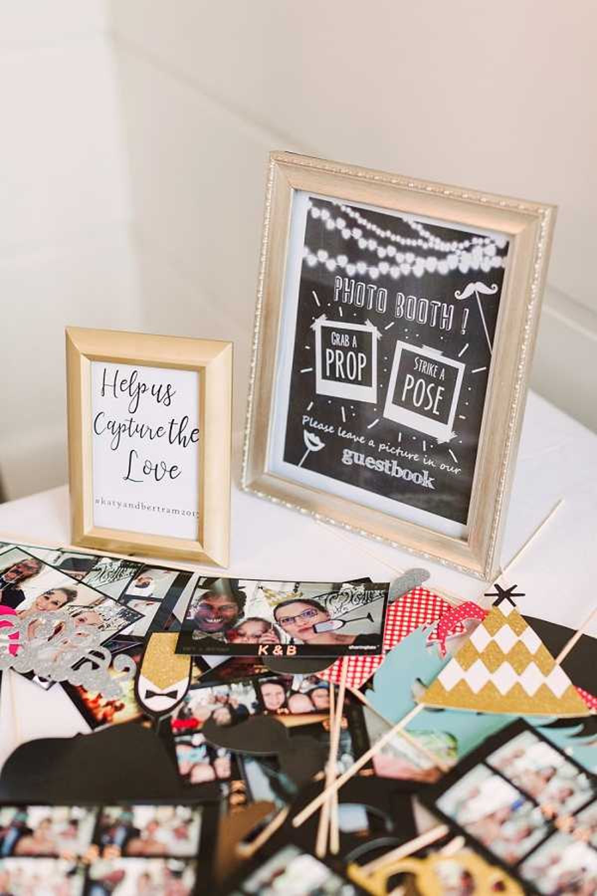 Detail shot of a wedding photo booth station set up on a white linen-covered table. Two framed signs are displayed: a small gold-framed white sign reading 'Help us Capture the Love #katyandbertram2017' and a larger silver-framed chalkboard-style sign reading 'Photo Booth! Grab a Prop, Strike a Pose – Please leave a picture in our guestbook.' The table surface is covered with printed photo booth strips showing guests posing, alongside a collection of props including gold glitter mustaches, a gold and white geometric pennant, a 'K & B' banner, and various black cardstock props on wooden sticks. The styling uses a black, white, and gold color palette with a modern, playful reception activity theme.