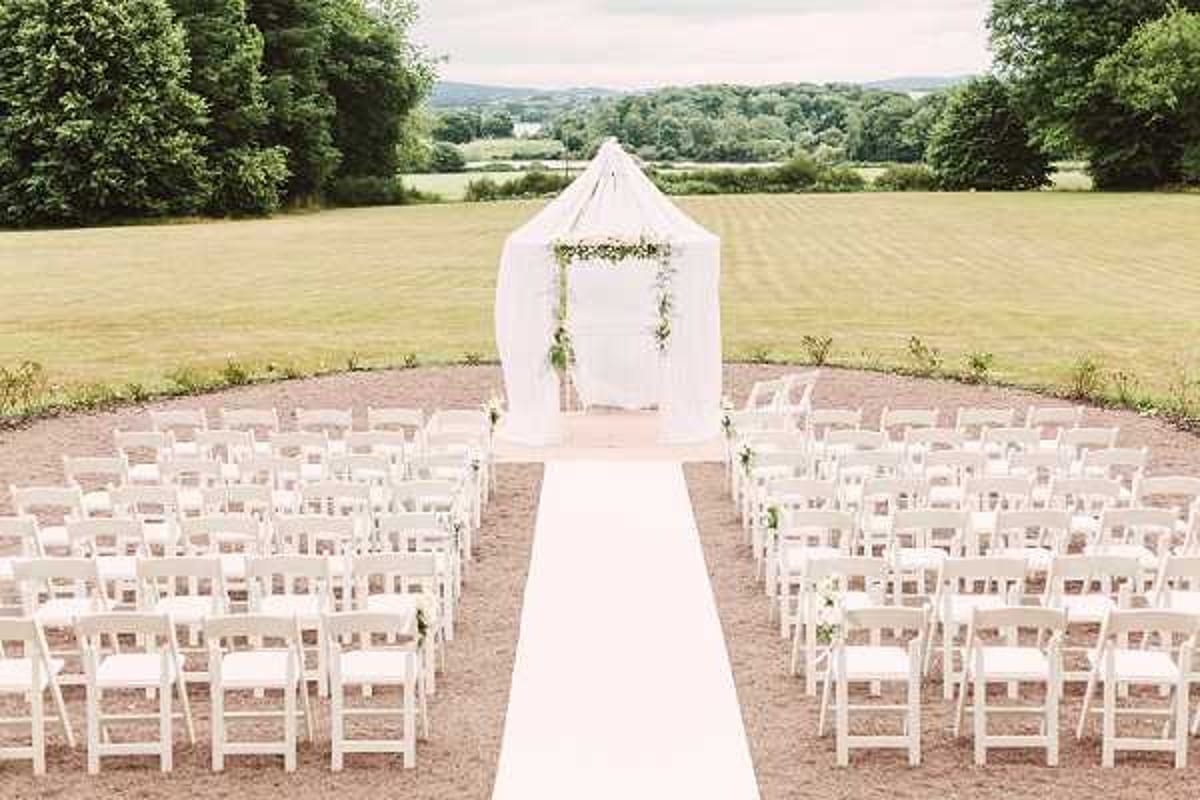 An outdoor wedding ceremony setup photographed from a slightly elevated wide-angle perspective, showing a symmetrical arrangement of white folding chairs divided into two sections by a white aisle runner. At the far end of the aisle stands a square arch decorated with greenery and small white florals, framed by a draped white fabric canopy structure. The ceremony space is set on a gravel or packed earth circular area, and no guests or wedding party are present, suggesting the photo was taken before the ceremony began. The overall decor palette is white and green, with a classic and clean styling aesthetic. Potential venue feature image.