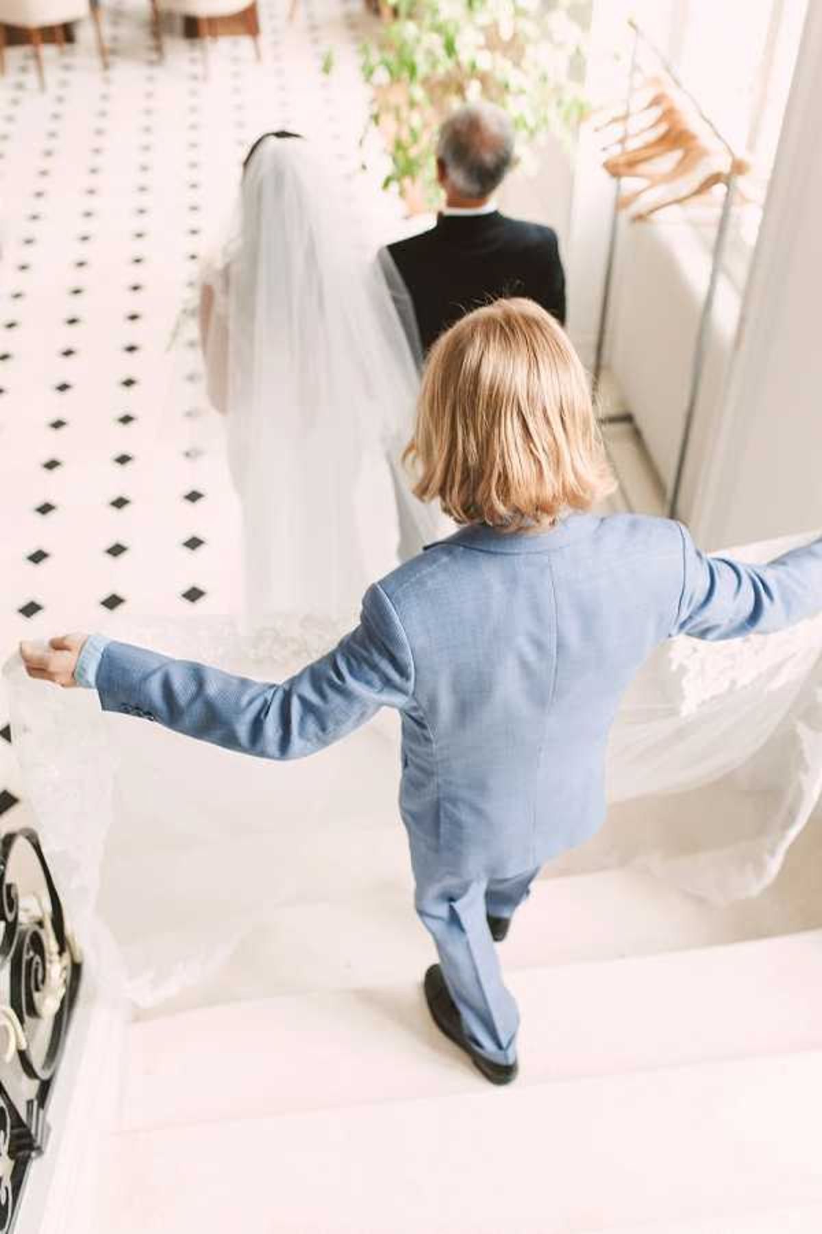 A getting-ready or pre-ceremony moment captured indoors, showing a child in a pale blue suit carrying the train of a bride's white gown as she walks ahead accompanied by an older man in a dark suit, likely a father figure. The setting appears to be an upscale interior with a classic black-and-white geometric diamond-patterned tile floor, white marble stairs with ornate black ironwork railing, and hanging garment racks visible near a bright window. The shot is taken from an elevated overhead angle looking down, giving a clear view of the veil and dress train being carefully held by the young attendant. The overall decor palette is crisp white and black with the child's dusty blue suit providing a color accent.