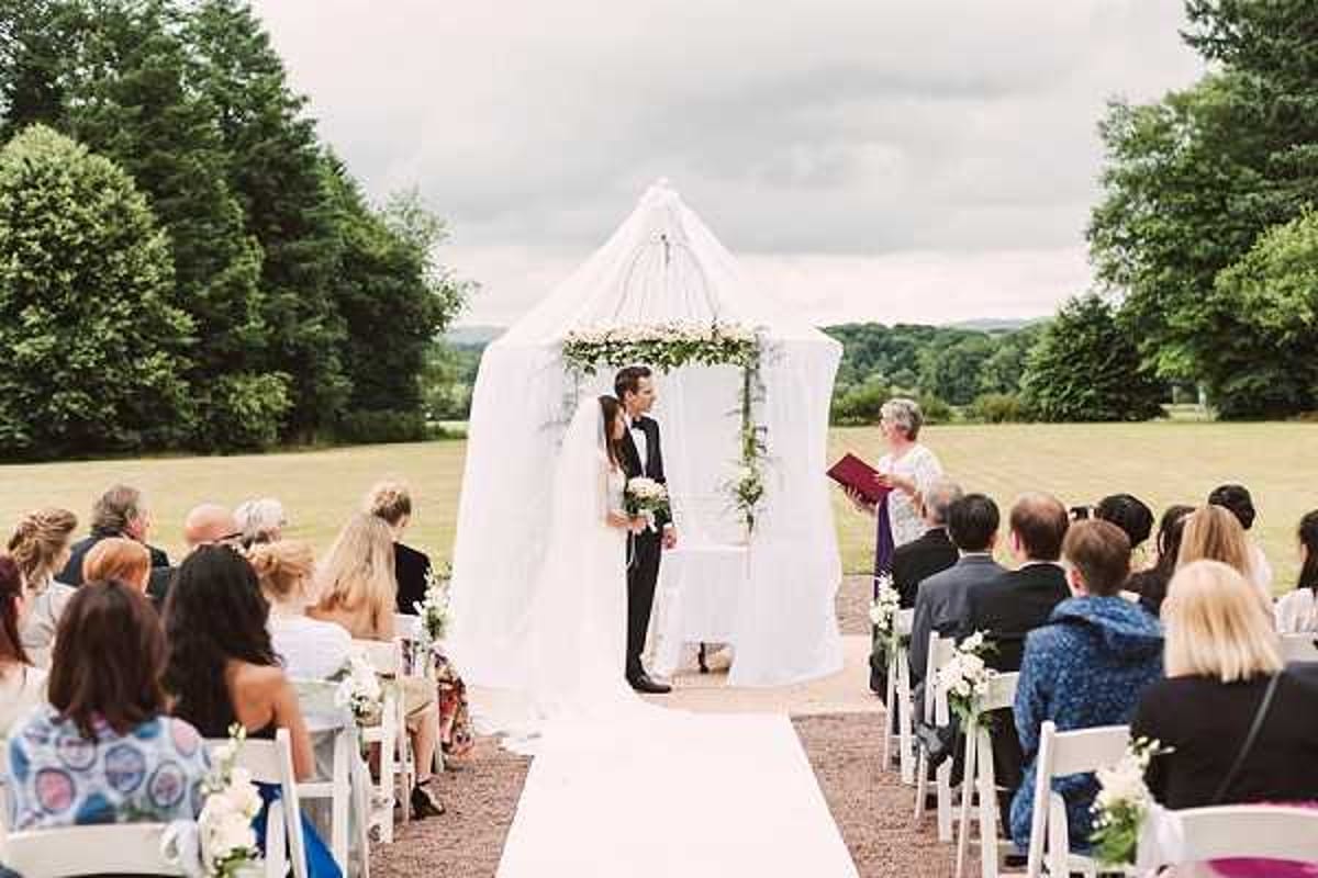 An outdoor wedding ceremony taking place on a large open lawn, with the couple standing beneath a white draped canopy structure adorned with a garland of white florals and greenery at the top. The bride wears a fitted white gown and holds a small bouquet of white flowers, while the groom is dressed in a black tuxedo with a white dress shirt. An officiant holding a red book stands facing the couple. Approximately 40-50 guests are seated on white folding chairs arranged in two sections with a white aisle runner between them, and small white floral arrangements are visible on the aisle chairs. The ceremony setup has a classic, formal style with an all-white decor palette. Wide shot taken from behind the seated guests, capturing the full ceremony scene.