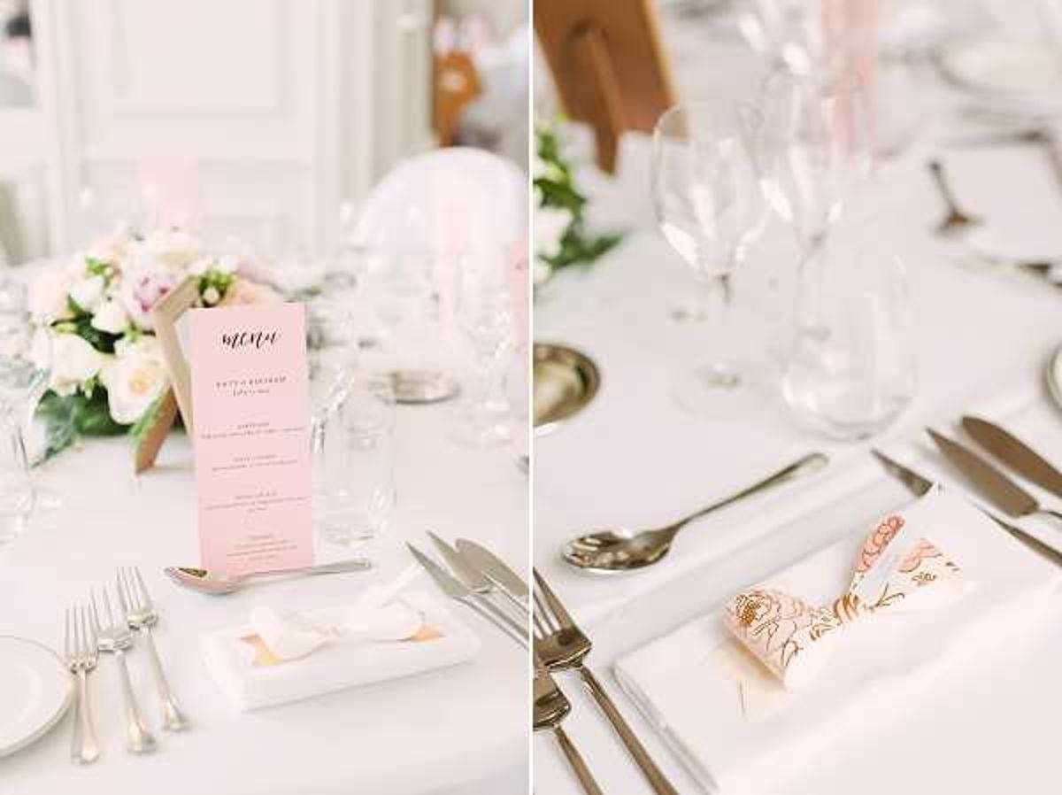 Close-up detail shots of wedding reception table settings in a bright, white interior venue. The left image shows a round table with a white linen, silver cutlery, crystal wine glasses, a blush pink printed menu card, a small white folded paper detail on a white square plate, and a low floral centerpiece featuring white and blush pink roses with greenery. The right image shows a close-up of a single place setting with a white linen napkin folded to hold a blush pink and gold floral-illustrated card, accompanied by silver cutlery and crystal glassware. The overall decor palette is white, blush pink, and gold with a classic, refined styling.