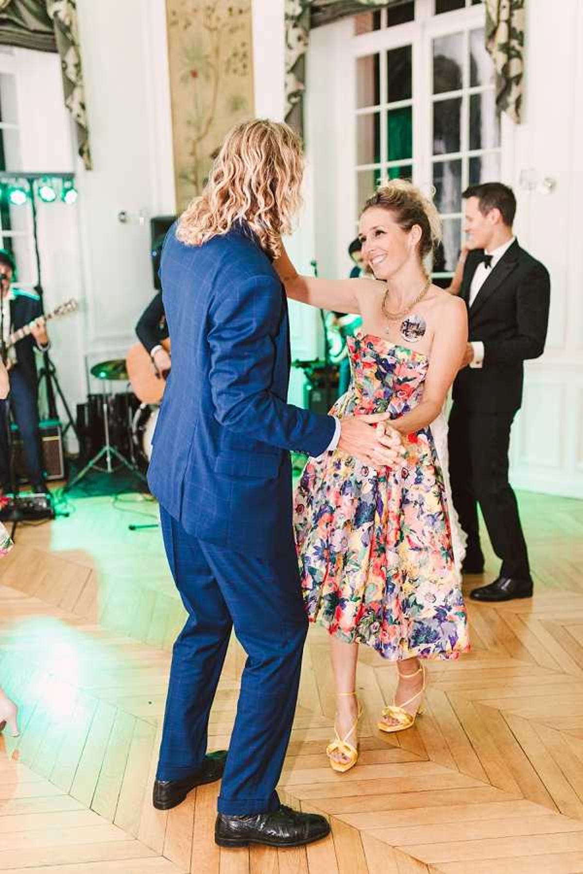 Two guests dance together on a parquet wood floor during the wedding reception in what appears to be an indoor ballroom or salon with ornate white paneling and large French windows. The man wears a cobalt blue suit with black shoes, and the woman wears a strapless midi-length dress with a multicolored floral print in pink, yellow, purple, and green, paired with yellow strappy heeled sandals. A live band performs in the background, with a guitarist visible, and another guest in a black tuxedo stands nearby. The setting has a classic French interior style, and the shot is taken at mid-range, capturing the dancers in full from a slight side angle.